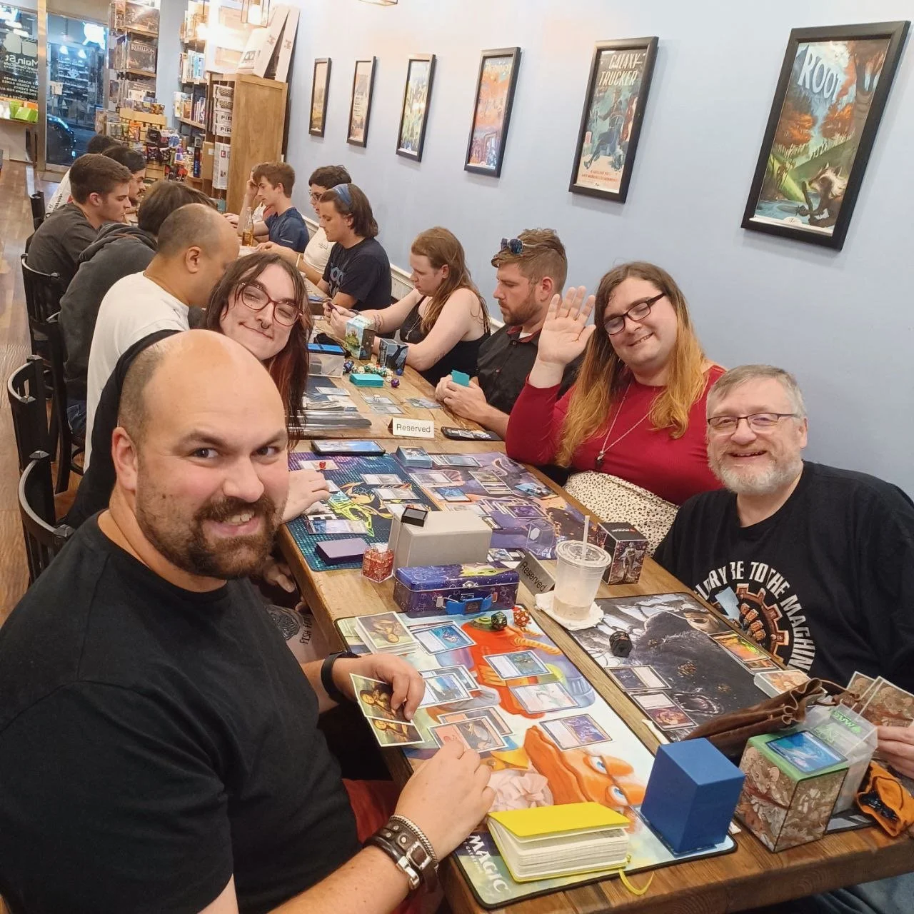 A group of people playing a tabletop card game at a long wooden table in a game store, with game artwork on the wall and various gaming accessories on the table.