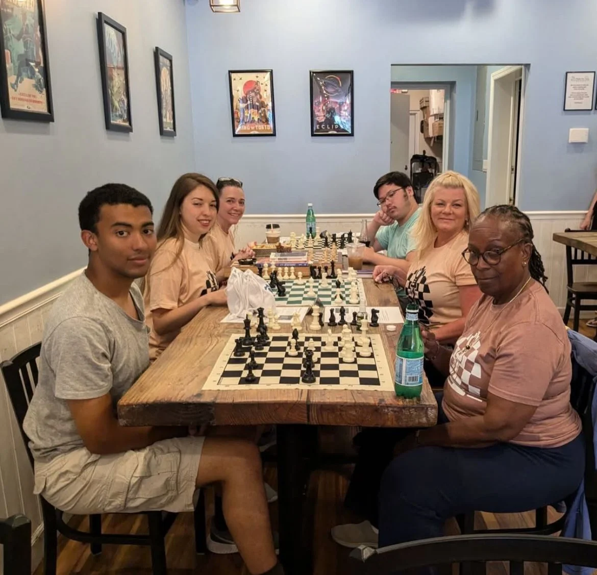 Group of six people playing chess at a wooden table in a café or restaurant with light blue walls and framed posters.