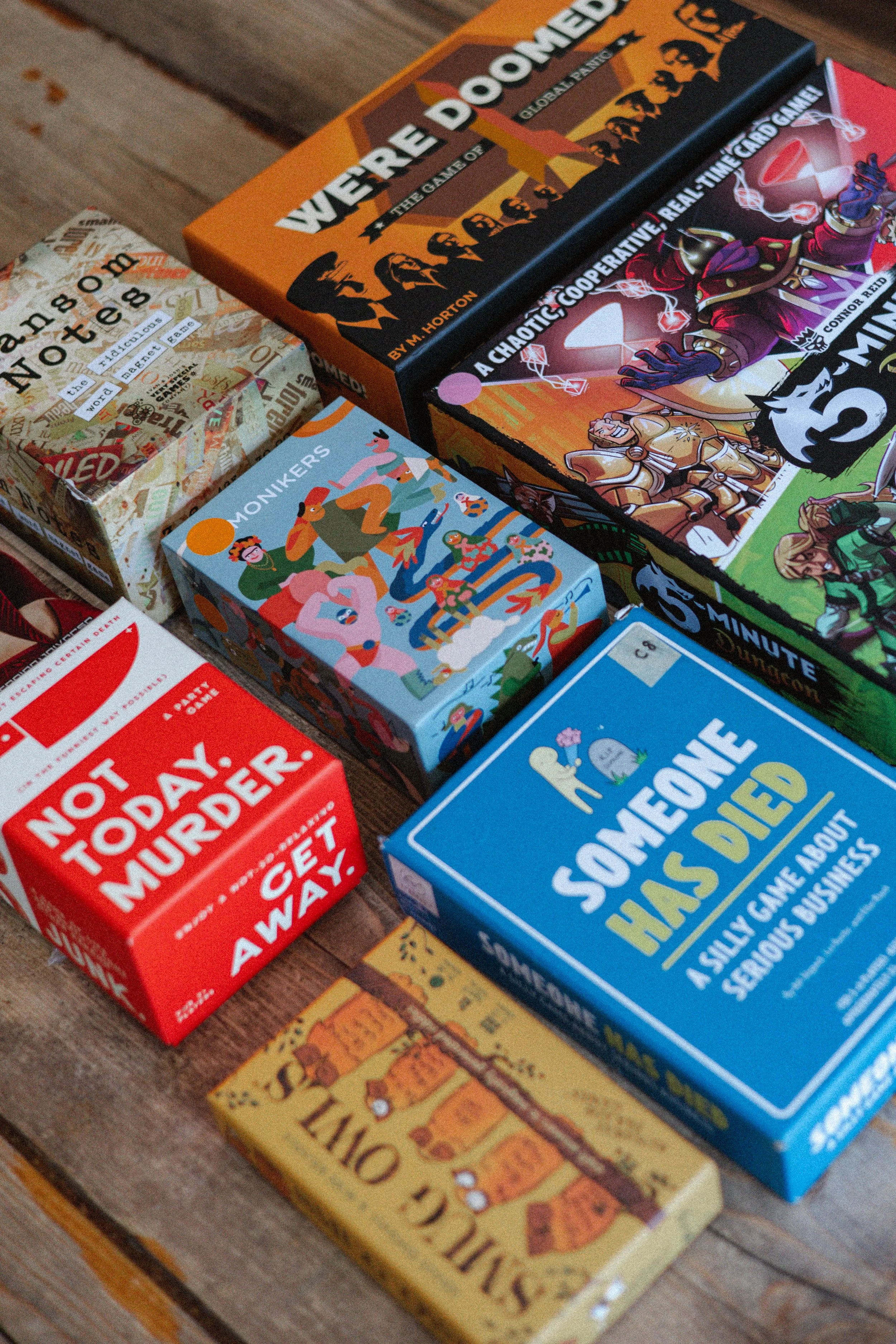 Photo of several tabletop board games, including 'We're All Doomed,' 'Someone Has Died,' 'Not Today, Murderer,' and others, arranged on a wooden surface.