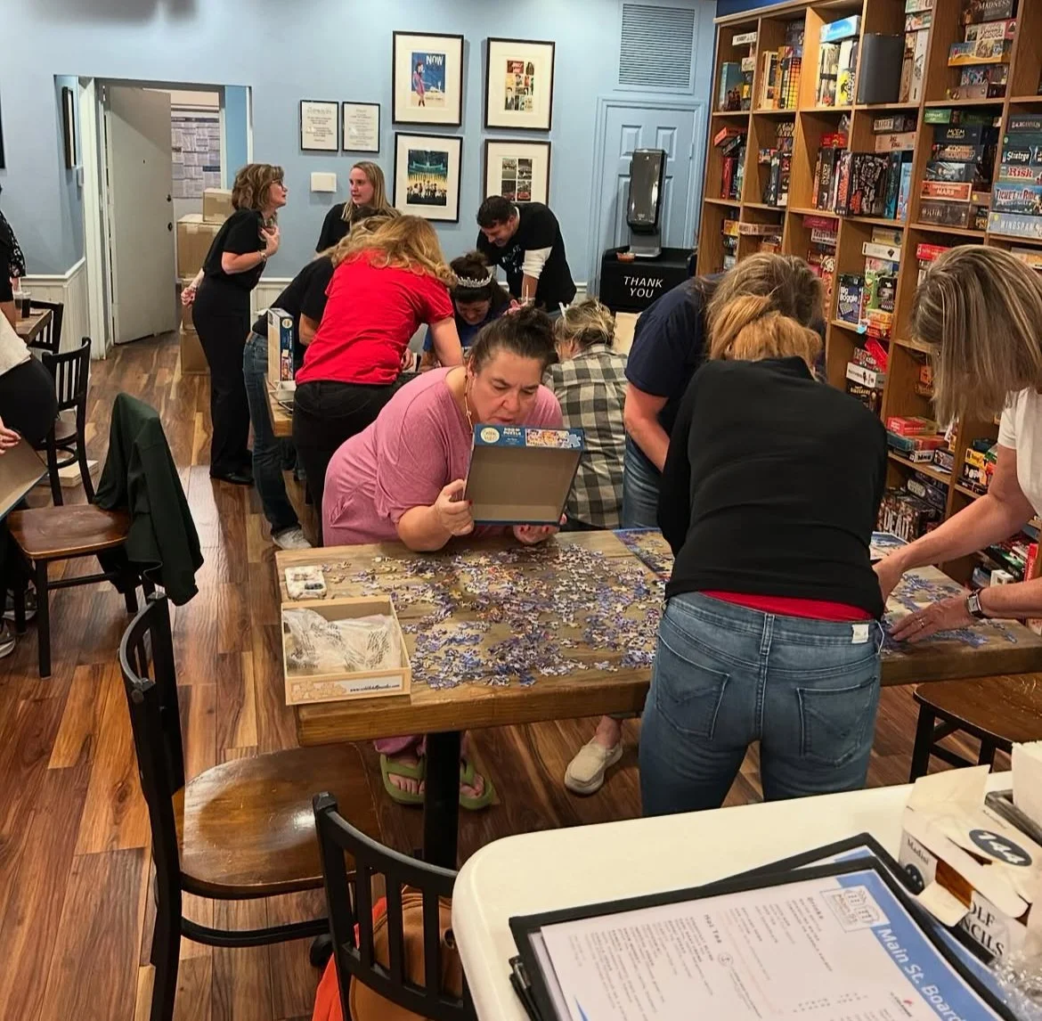 Group of people gathered around a wooden table working on a large jigsaw puzzle in the cafe with shelves of board games and framed pictures on the wall.