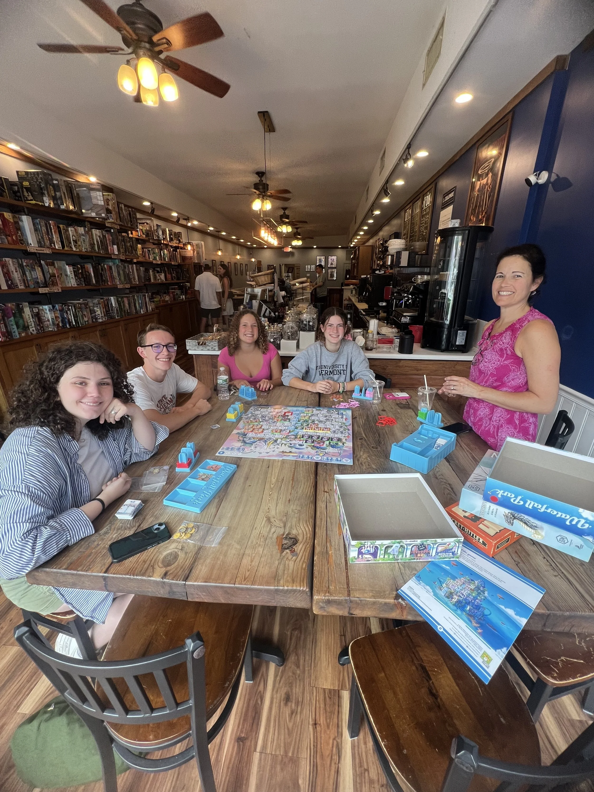 A group of five young women with one of the women standing, playing a board game at a wooden table inside the cafe, with bookshelves and a counter in the background.