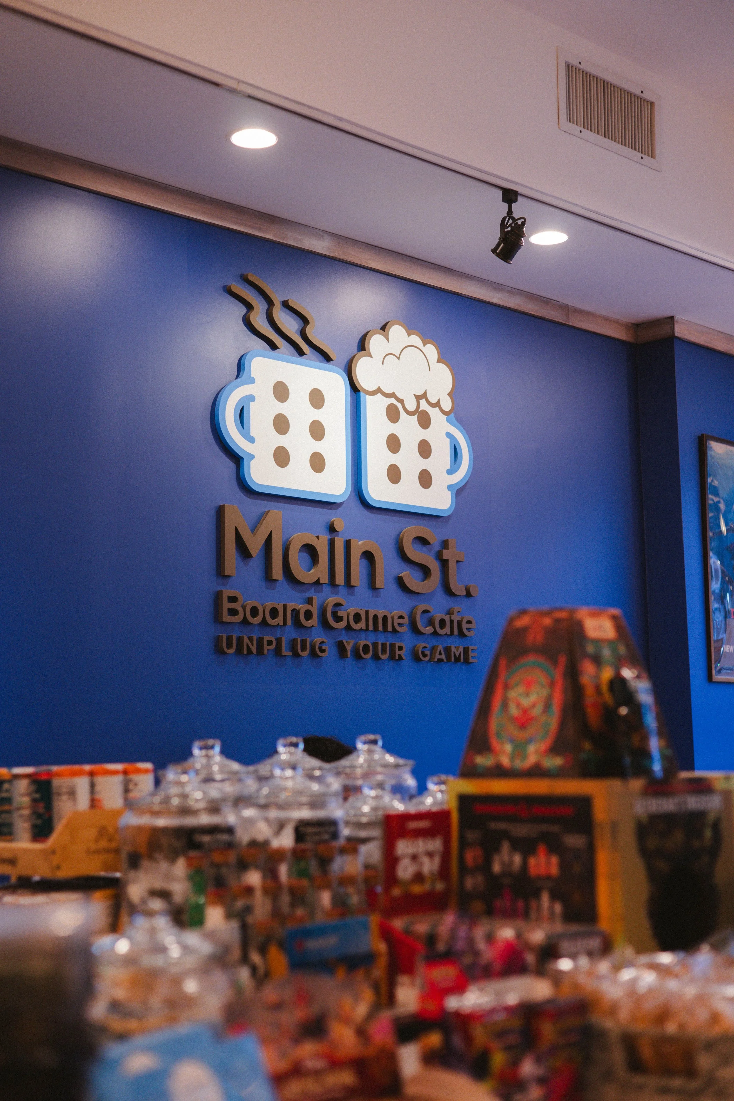 Interior of a board game cafe called 'Main St.' with a blue wall, a logo featuring two steaming beer mugs, and a tabletop with various board games and snacks.