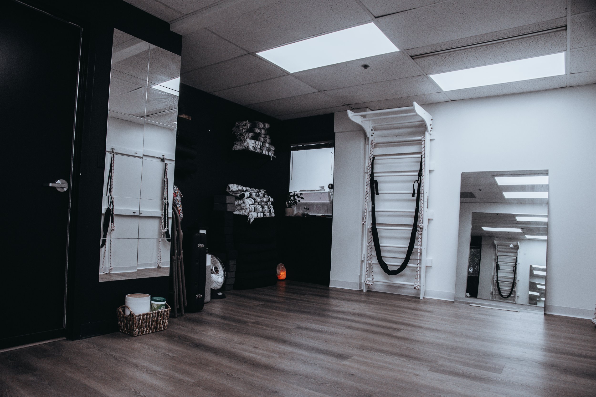 Interior view of a fitness or Pilates studio with black and white walls, neatly folded towels on shelves, a large mirror reflecting part of the room, fitness equipment including a wall-mounted ladder with resistance bands, and a wooden floor.