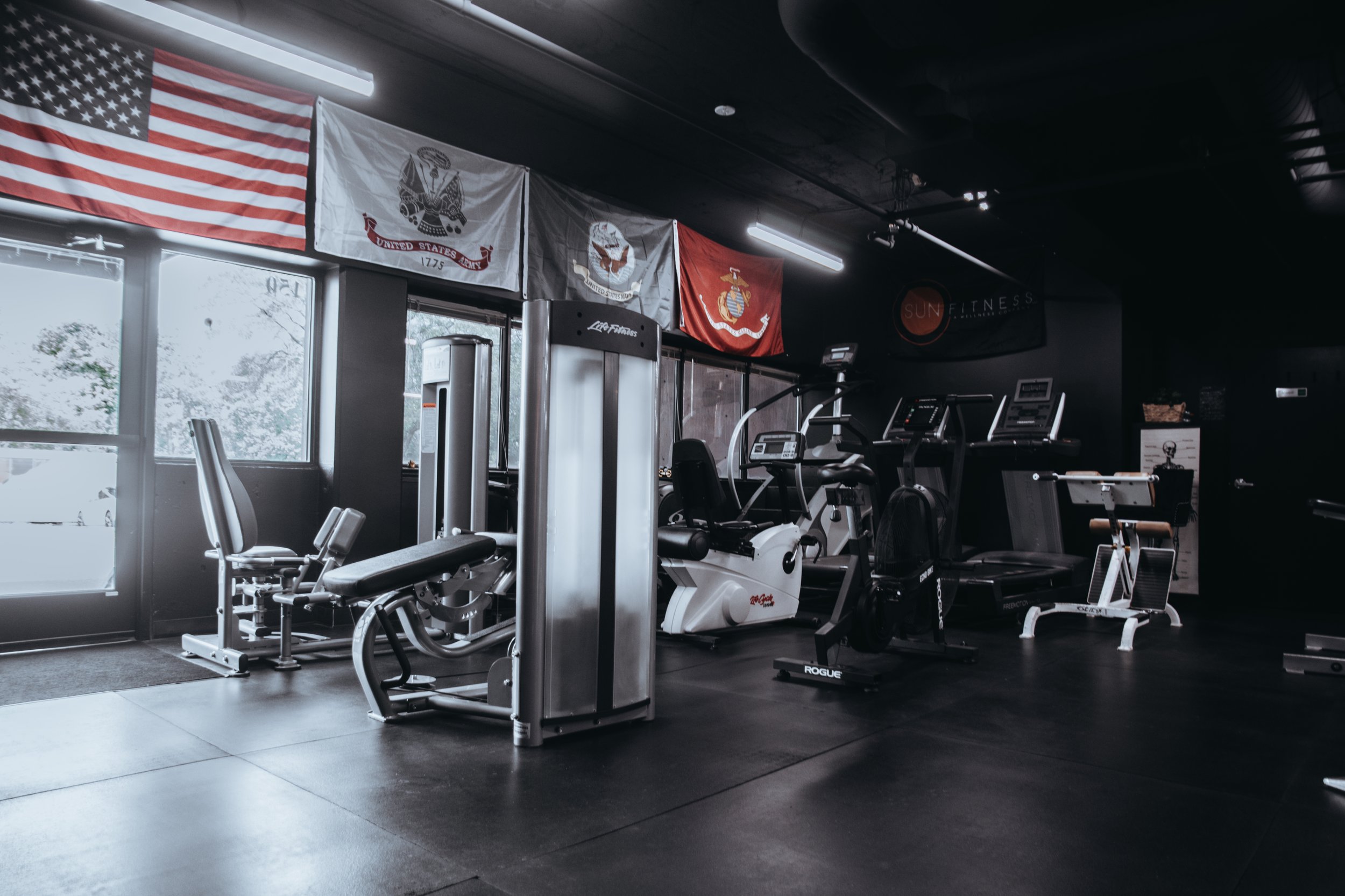 Empty gym interior with exercise equipment, military flags on wall, and large window letting in natural light.