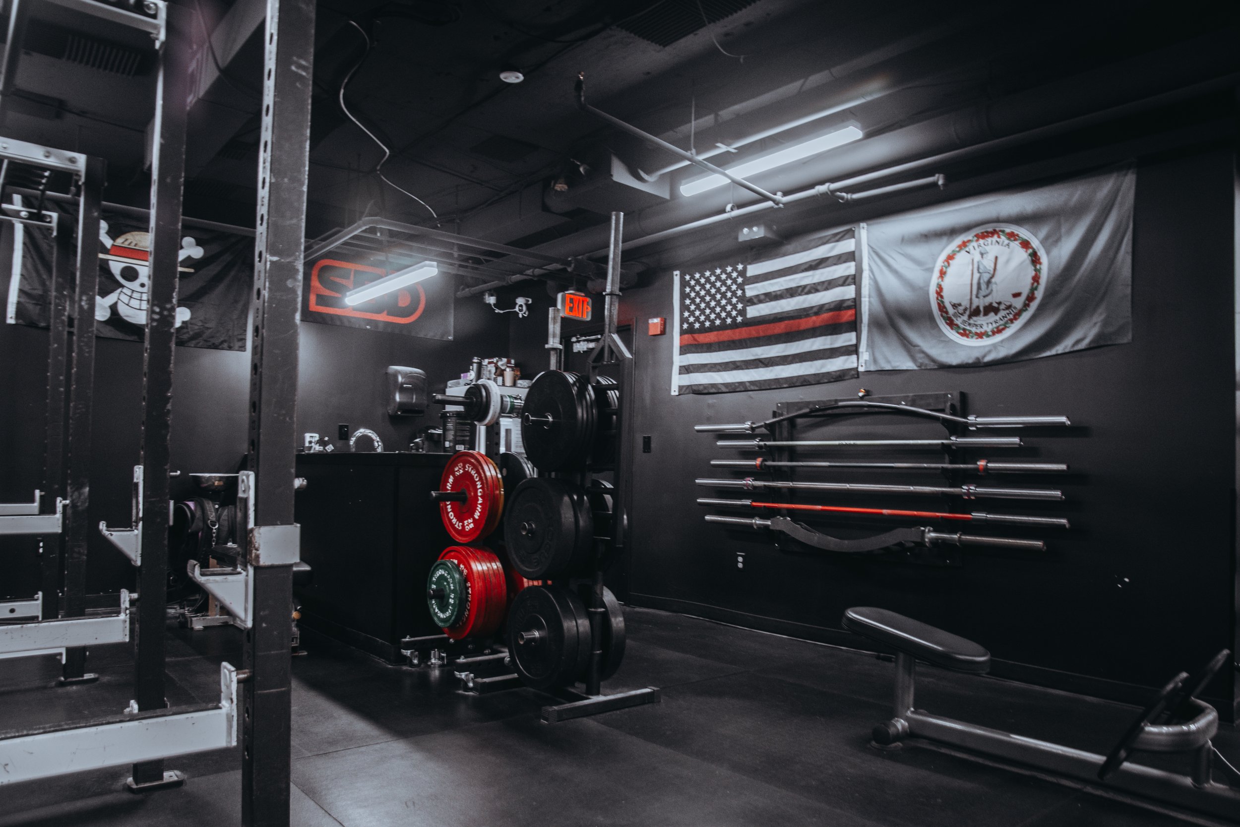A gym with black walls decorated with flags, weightlifting equipment, and a bench.