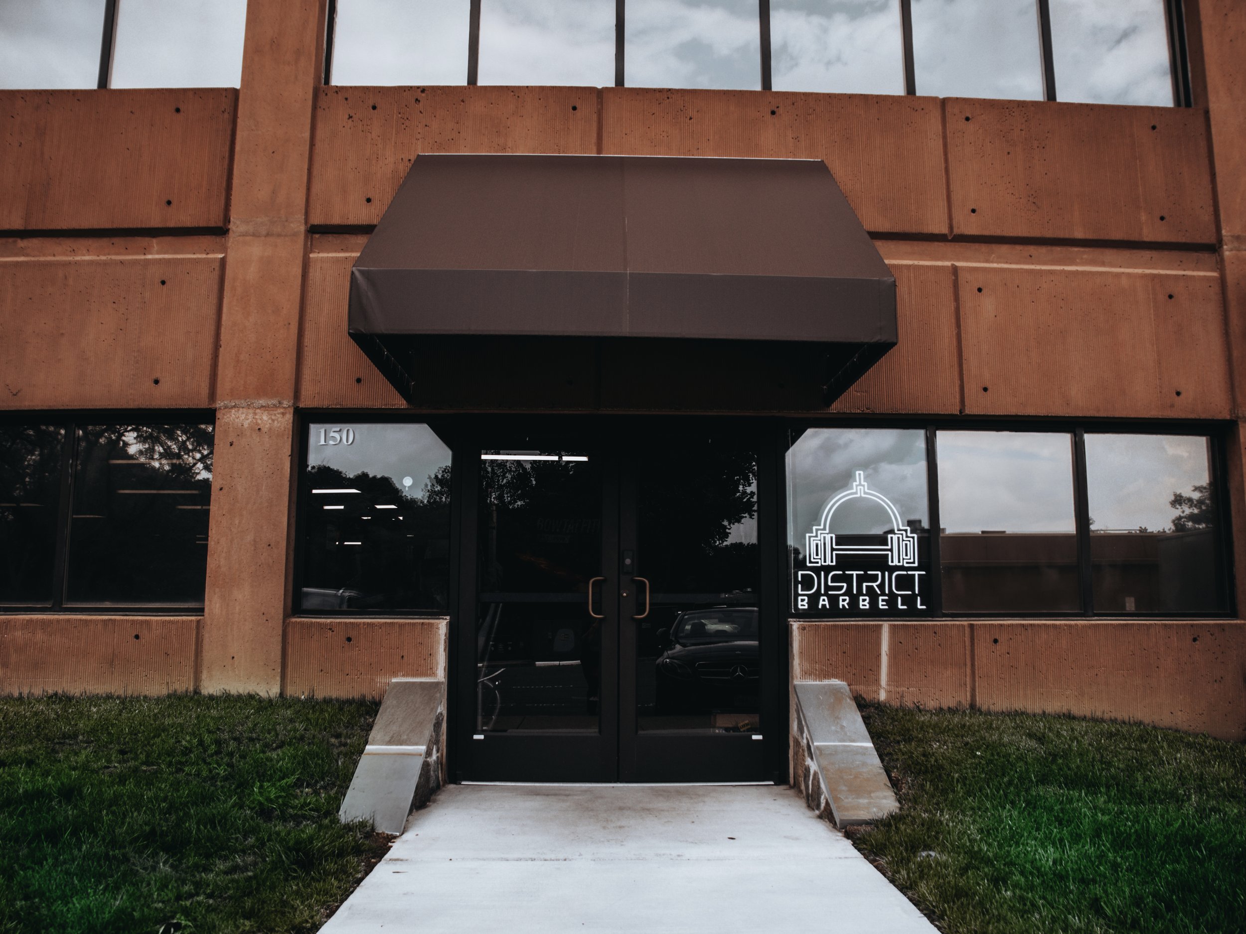 Exterior view of District Barbell gym with a black awning, glass doors, and a neon sign of a barbell on the window.