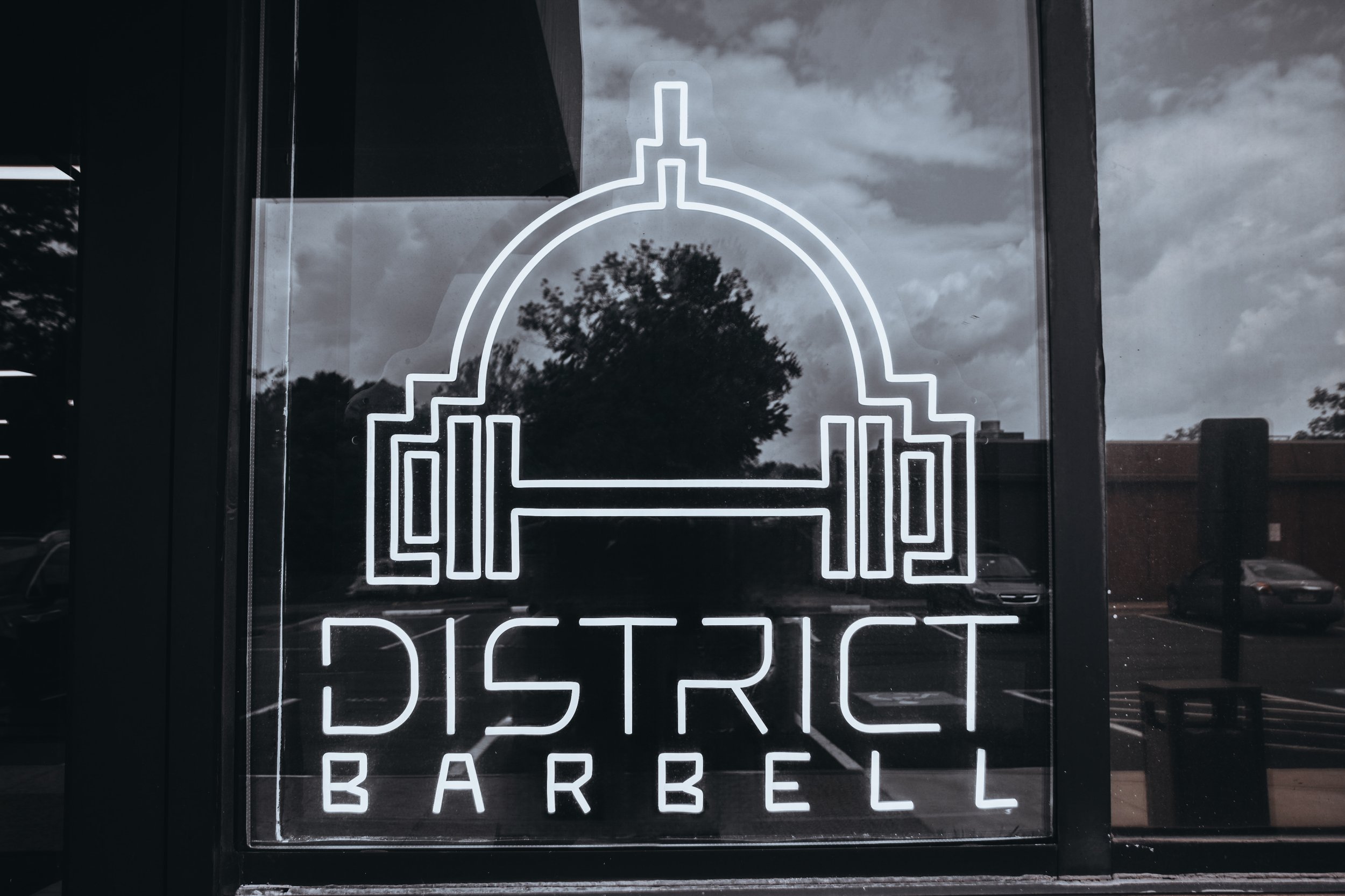 A neon sign with a stylized gym logo, featuring a barbell and the words "District Barbell" on a glass window reflecting trees and clouds.