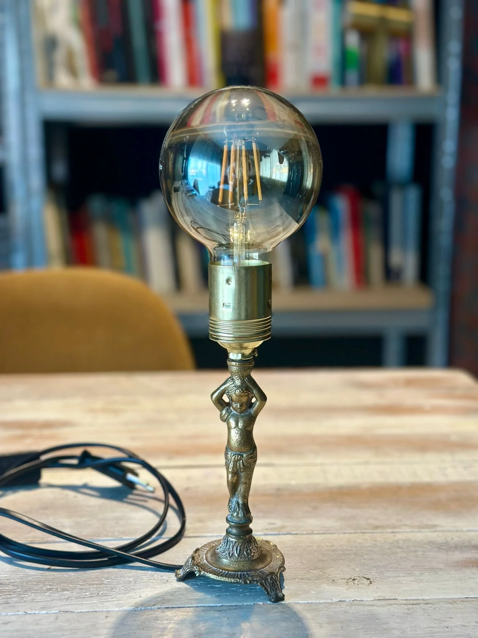 Lámpara de escritorio con base metálica y bombilla expuesta sobre mesa de madera, fondo de libreros con libros.