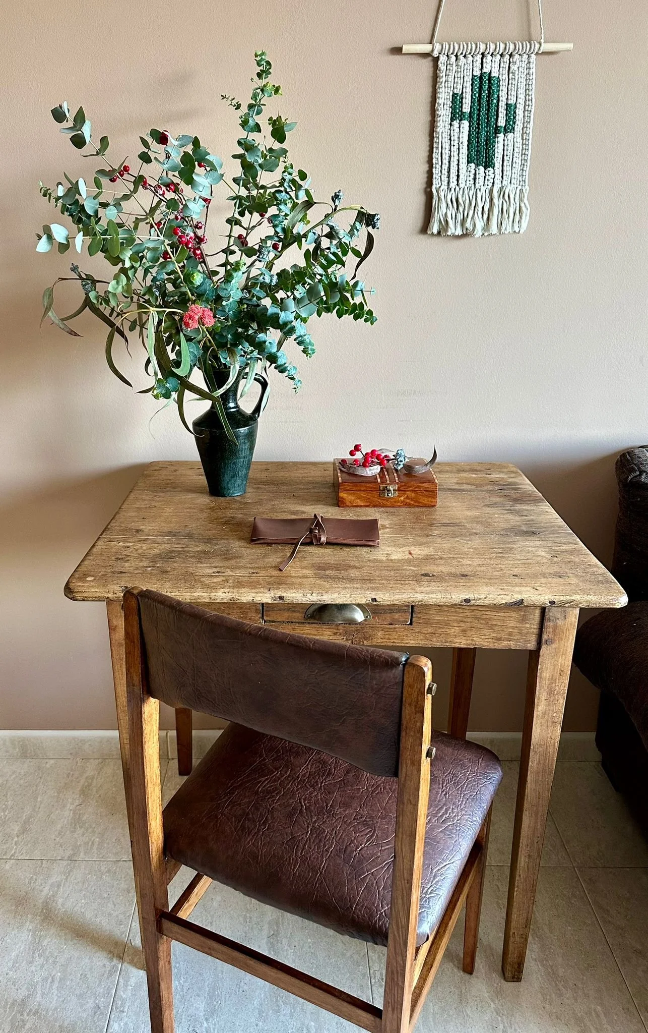 Mesa de madera rústica con una silla de respaldo alto de cuero marrón, un florero con plantas verdes y rojas, caja de madera con adornos y una cartera marrón. En la pared, un tapiz decorativo con símbolo de cruz verde.