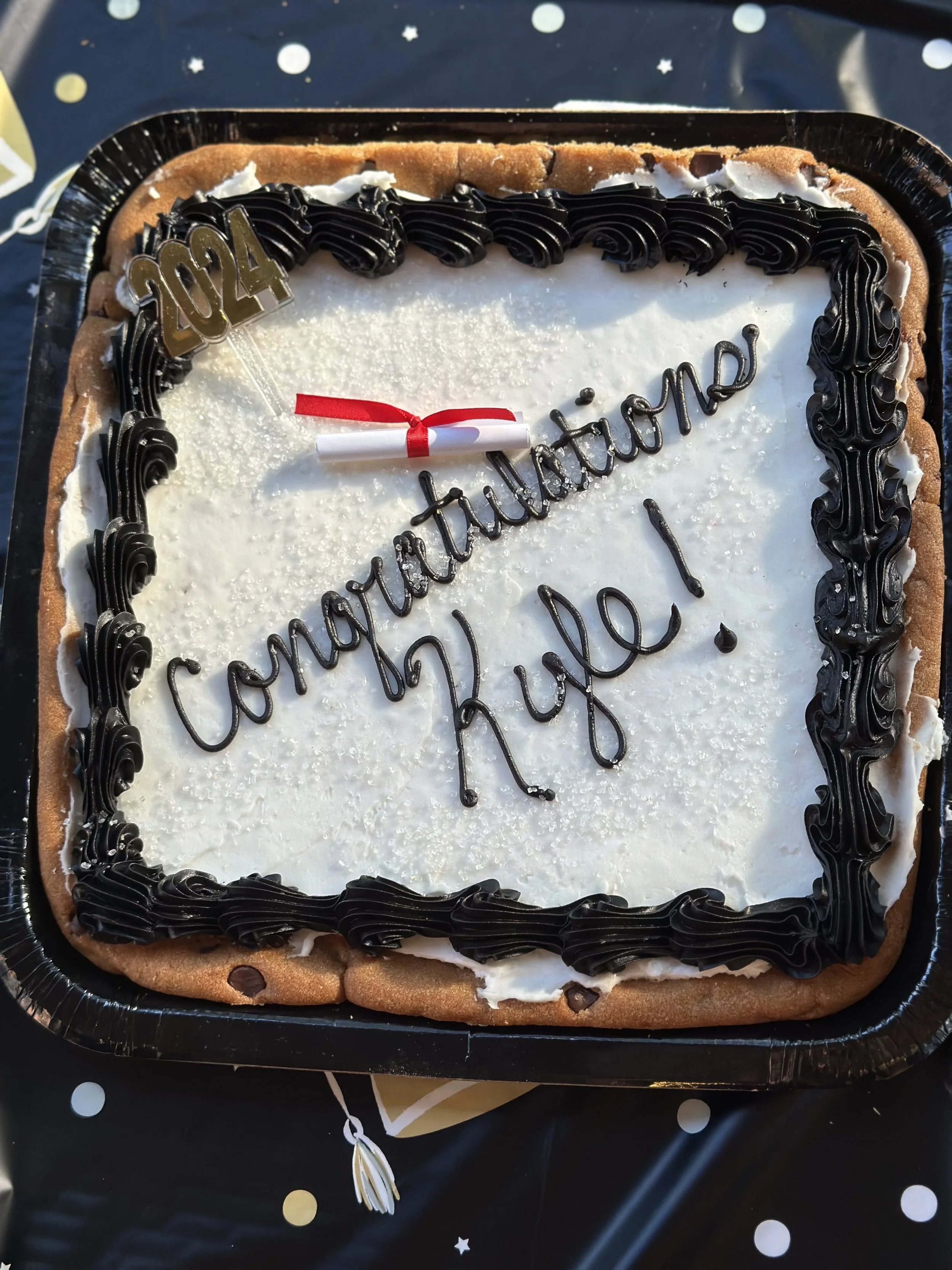 Square celebration cake with white frosting, black icing border, and a small rolled document with a red ribbon on top. The cake has a message written in black icing: 'Congraduations! Kuge!' and a '2024' gold decoration on the upper left corner.