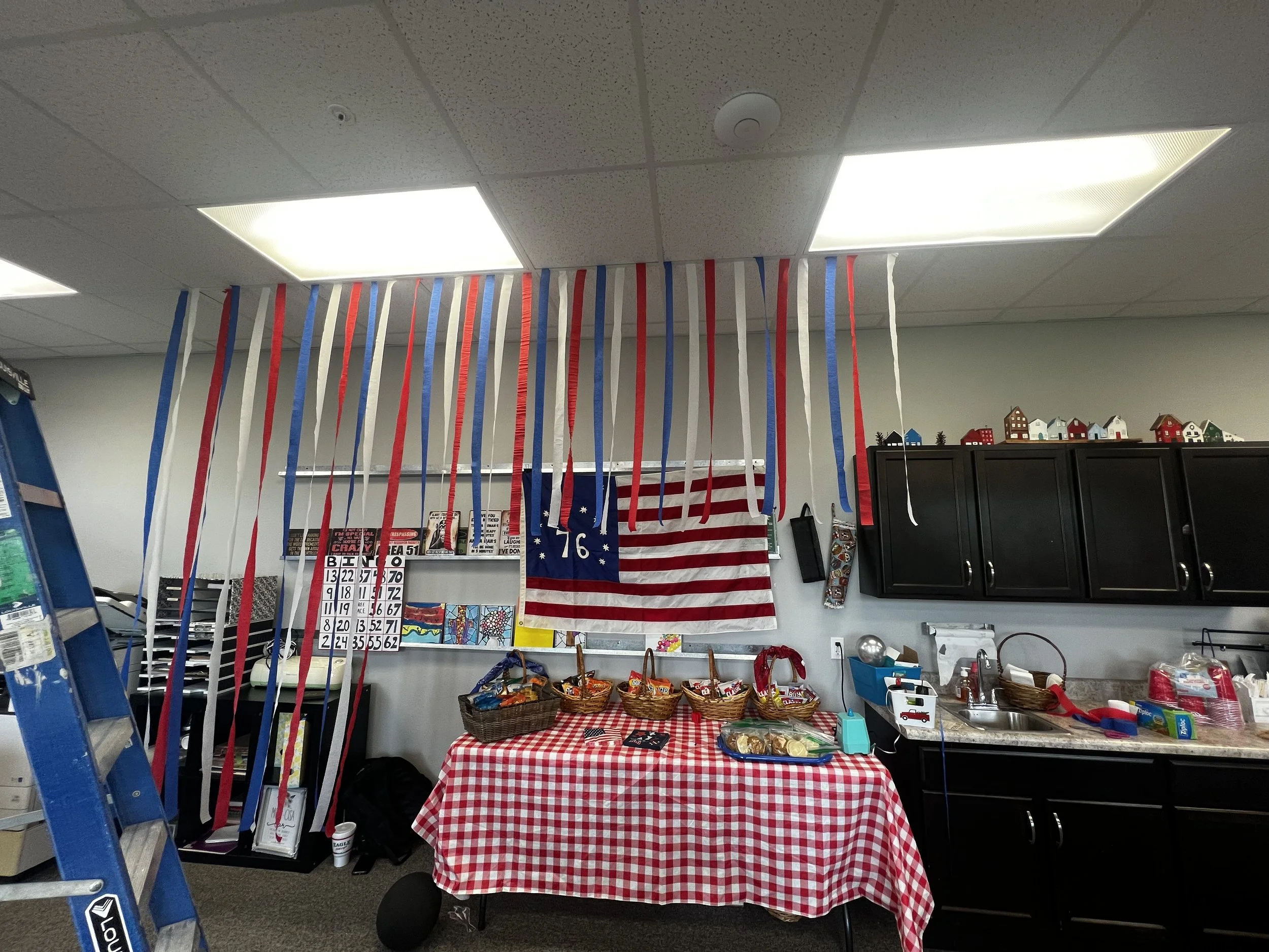 Decorative setup with red, white, and blue streamers hanging from the ceiling, American flags, and patriotic-themed banners and decorations, suggesting a celebration of a national holiday like Independence Day. The room also has a table with a red and white checkered tablecloth, baskets of baked goods, and various party supplies.