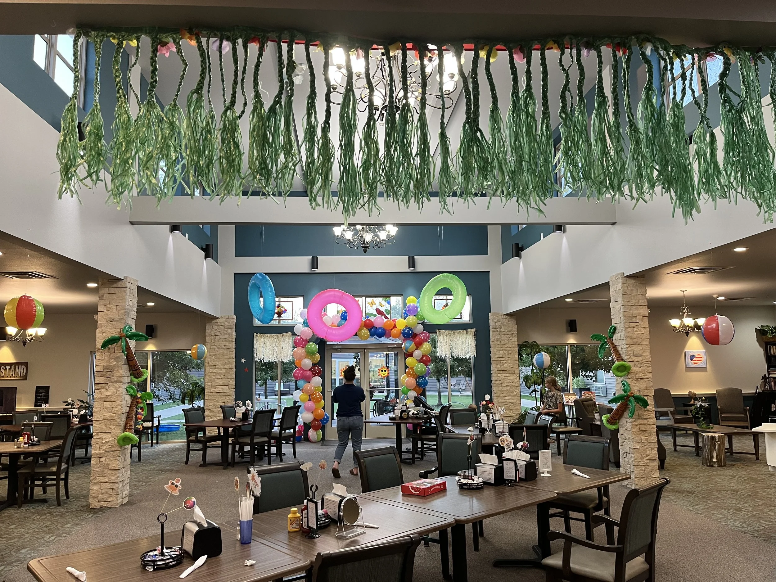 Decorated indoor space with balloons and hanging decorations for a party. Green paper strands hang from the ceiling, colorful balloon arch with rings and tropical-themed inflatables, tables set with condiments and decorations, and a few people present.