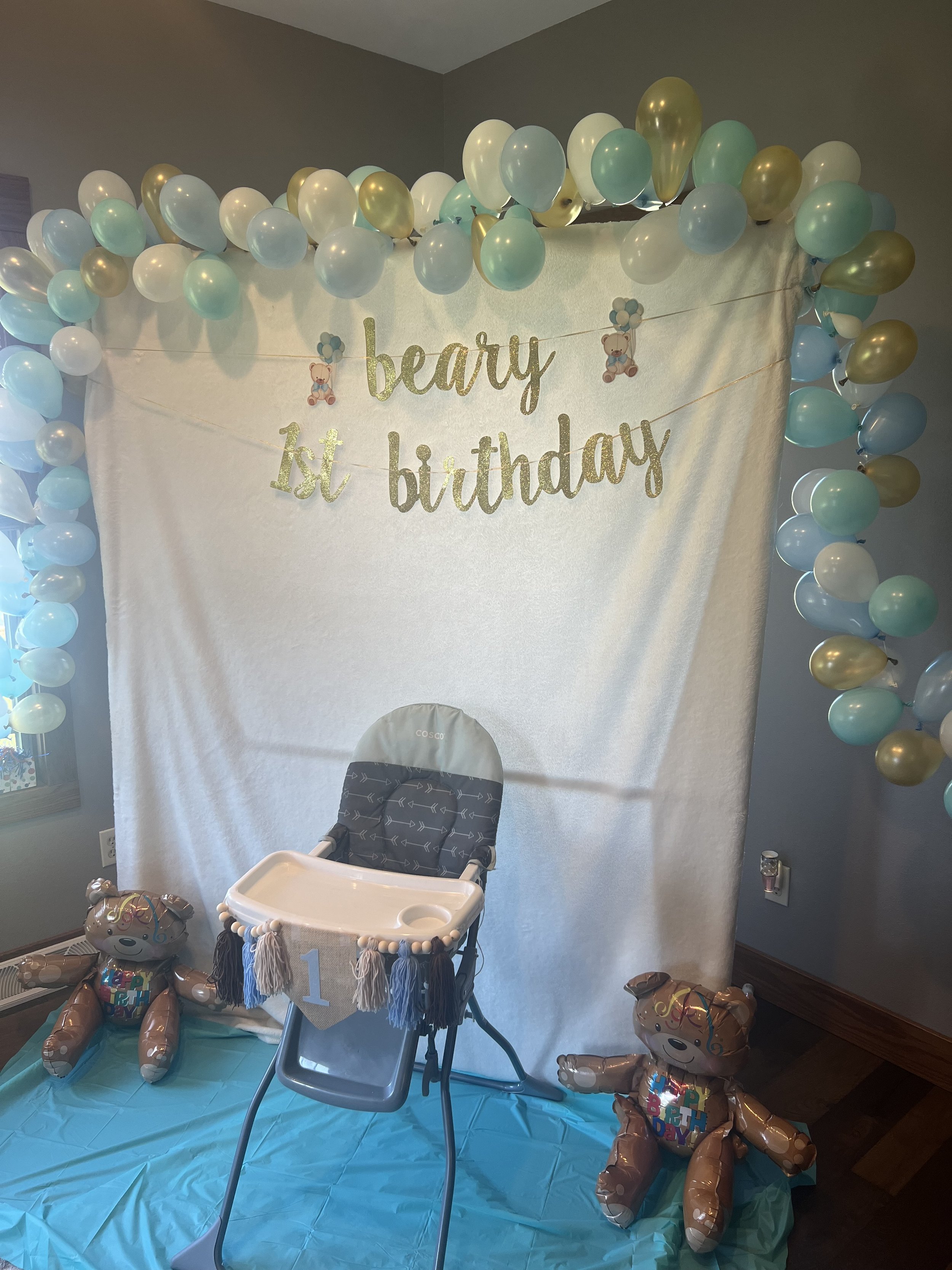 Decorated area with a white backdrop, gold and blue balloons, a banner that says 'beary 1st birthday,' a high chair with a small banner reading '1,' and two teddy bear balloons at the bottom.