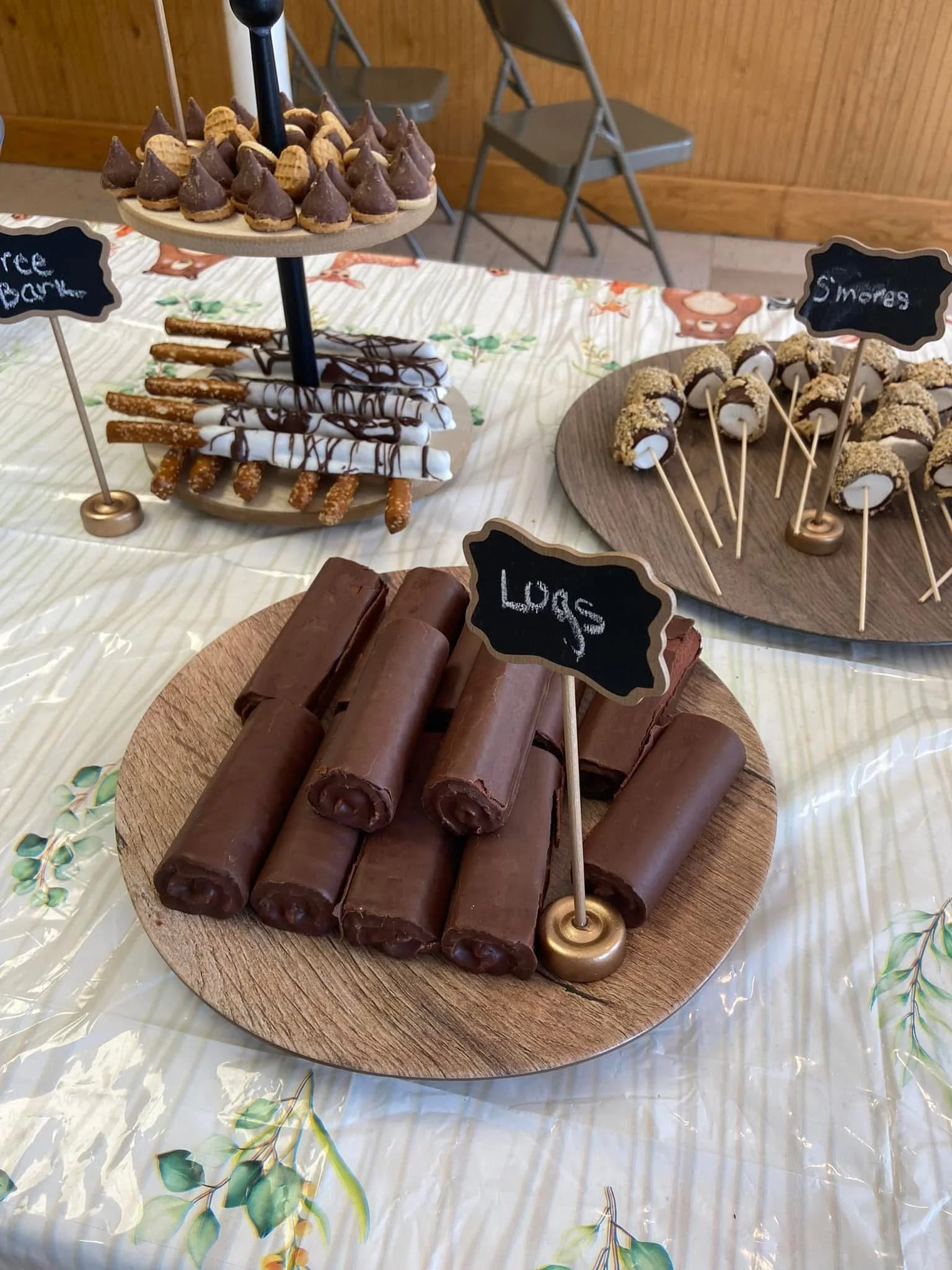 Chocolate logs on a wooden plate with a small chalkboard sign labeled 'Logs' in the foreground, with other dessert trays featuring chocolate-coated treats in the background.