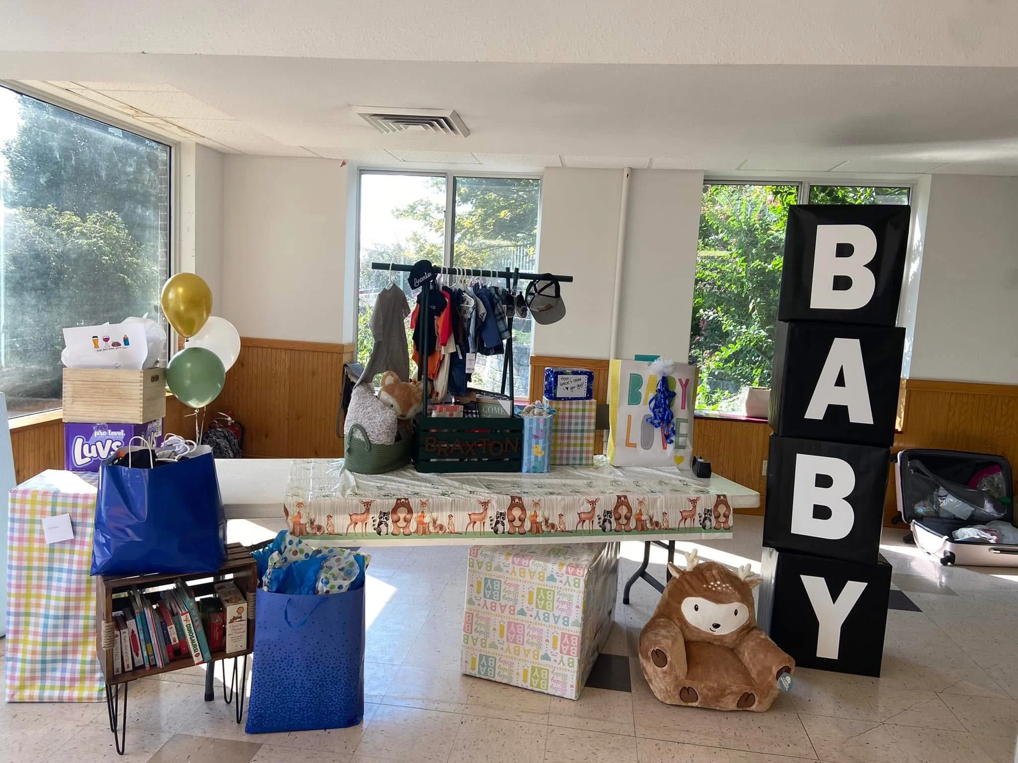 Indoor space decorated for a baby shower with a large black block sign spelling 'BABY', a table with gifts and decorations, balloons, and gifts including books and toys.