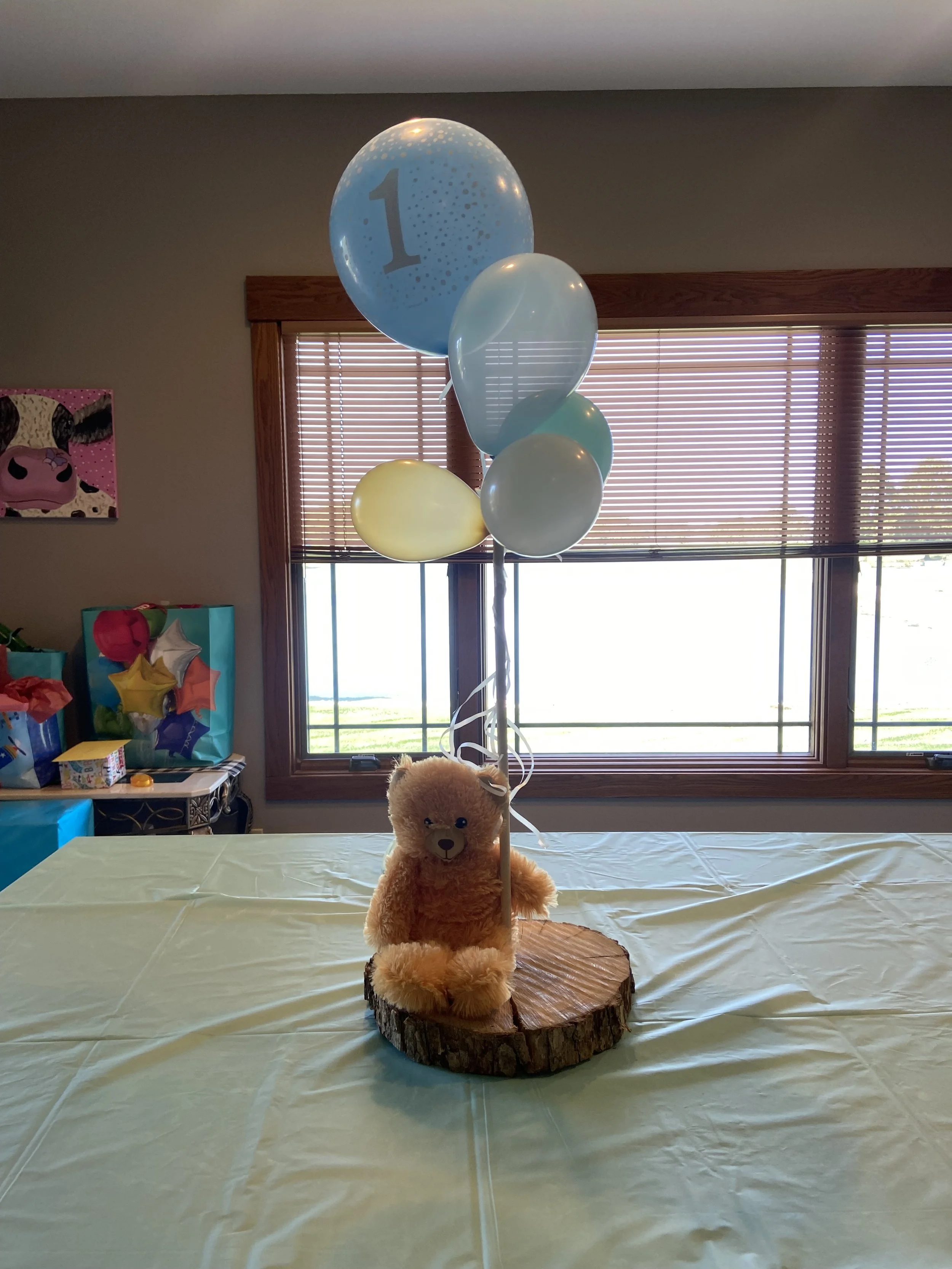 A teddy bear plush toy is sitting on a wooden round slab with four balloons attached to a stick next to it, including a blue balloon with the number 1 on it, indicating a first birthday celebration, on a table with a light-colored tablecloth. The background includes a window with blinds and colorful gift bags.
