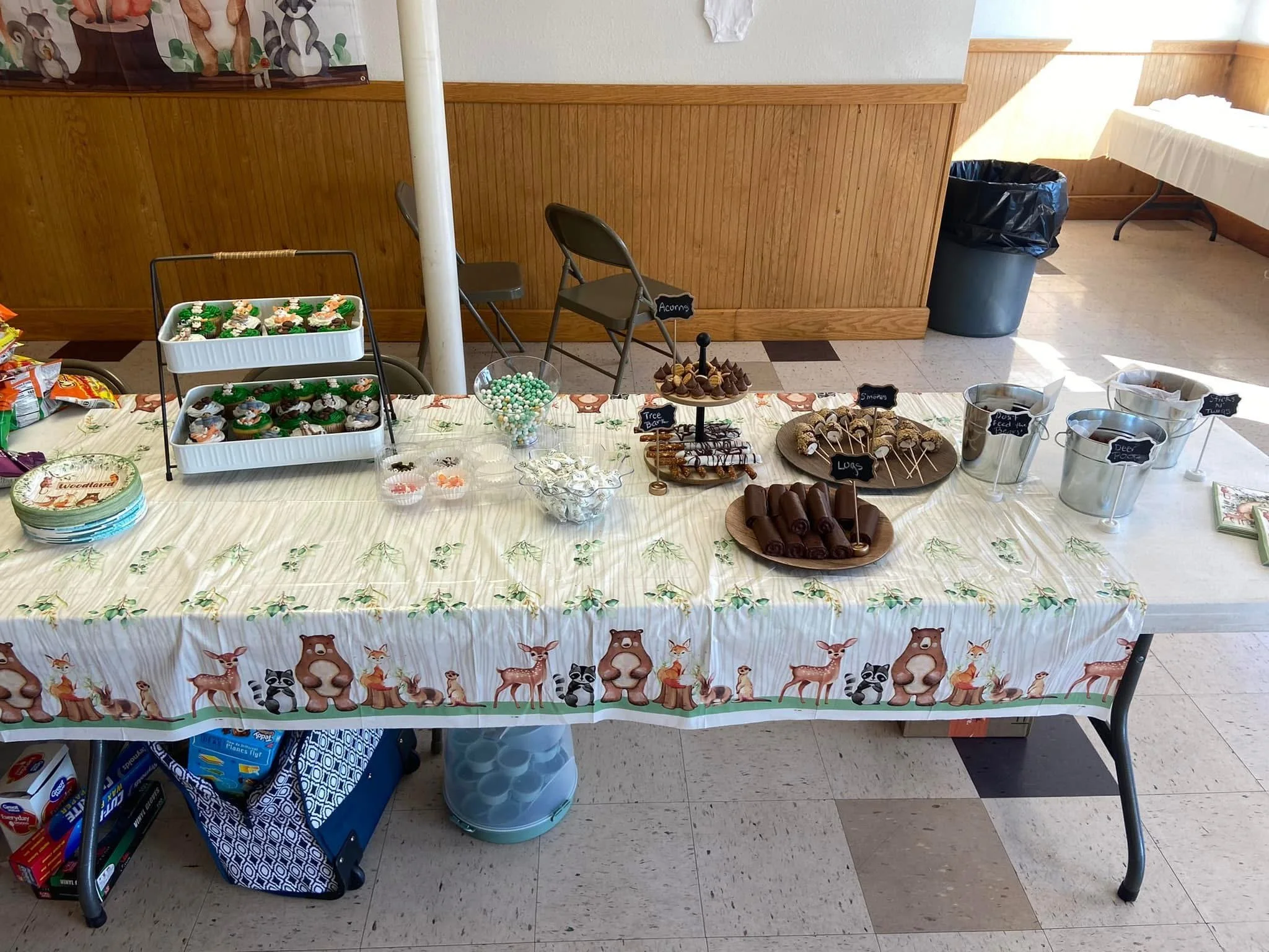 A table decorated with a holiday-themed tablecloth featuring reindeer and woodland animals, displaying various desserts and candies, including cupcakes with Christmas decorations, bags of chips, bowl of peppermint candies, and other sweets, set in a room with wooden paneling and folding chairs.
