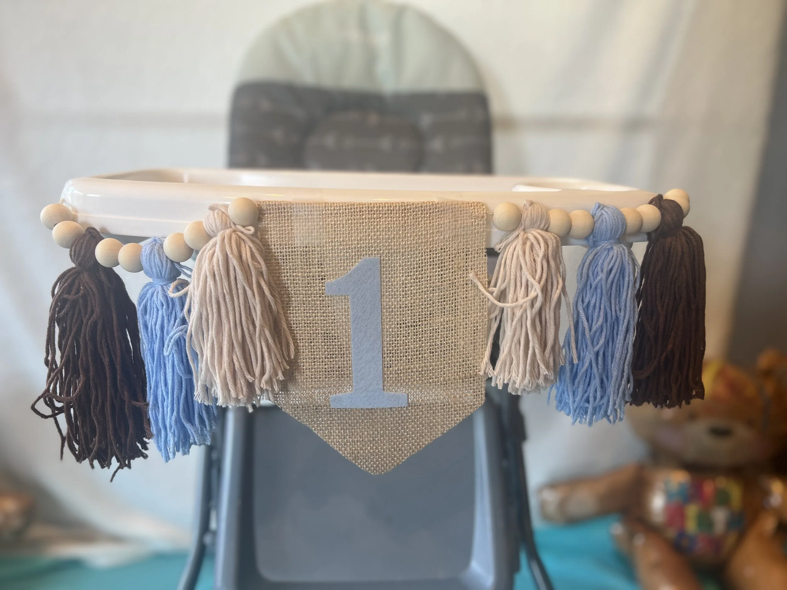 Decorative birthday banner with a beige fabric background, white number '1', and tassels in white, blue, and brown hanging from a white circular frame.
