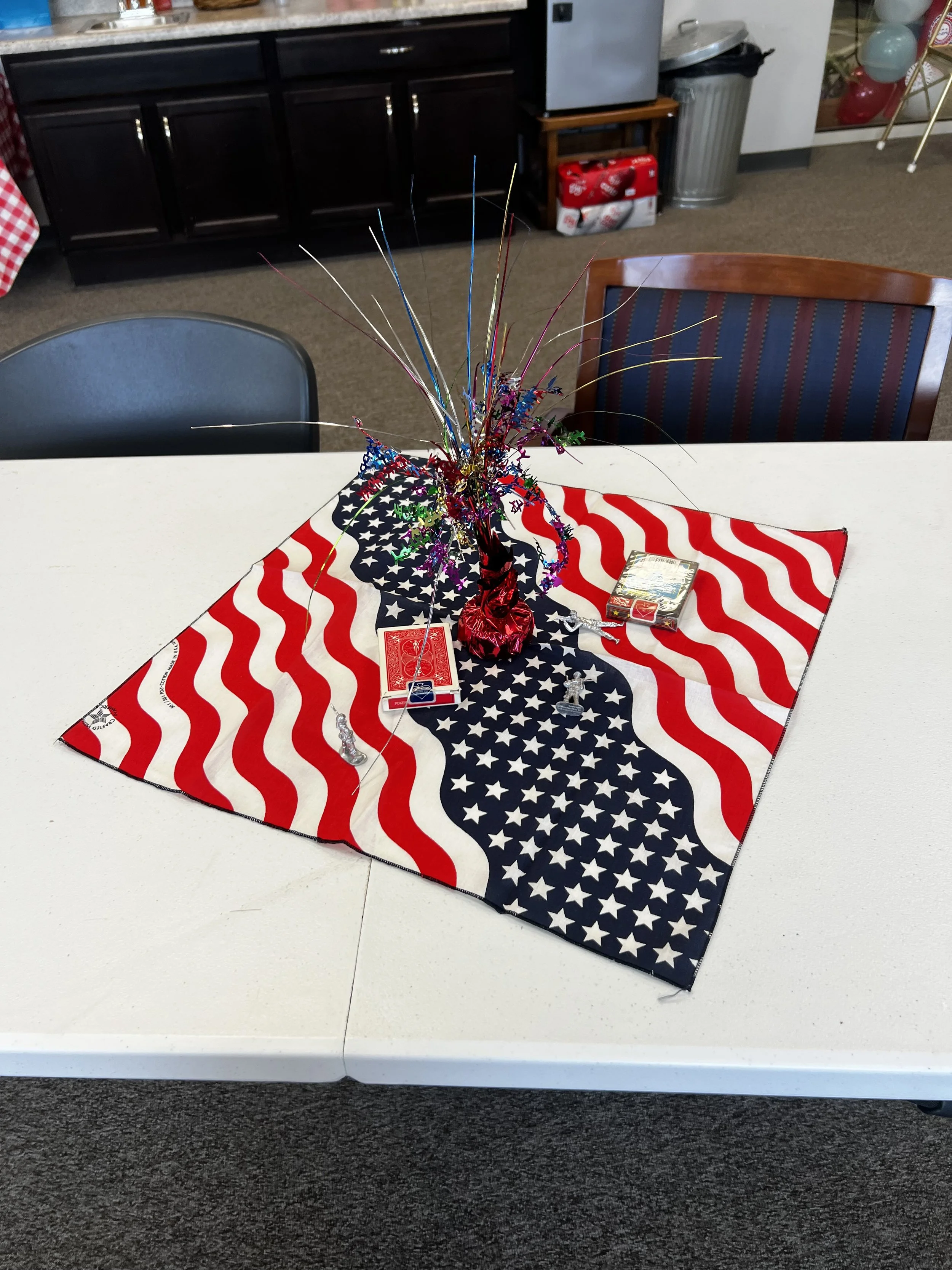 Table decorated with a star-spangled tablecloth, a patriotic centerpiece with multicolored tinsel and metallic wires, a deck of playing cards, and small decorative figures, in a room with black cabinets and holiday items in the background.