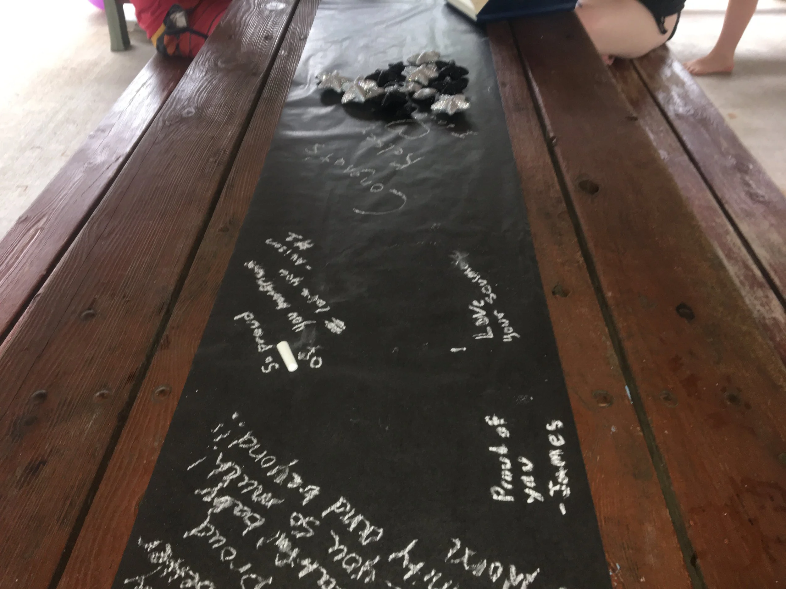 A black chalkboard table runner with handwritten food menu items in white chalk, surrounded by a wooden picnic table with people sitting around it, some with bare feet, and a small pile of foil-wrapped candies or chocolates at the top of the chalkboard.
