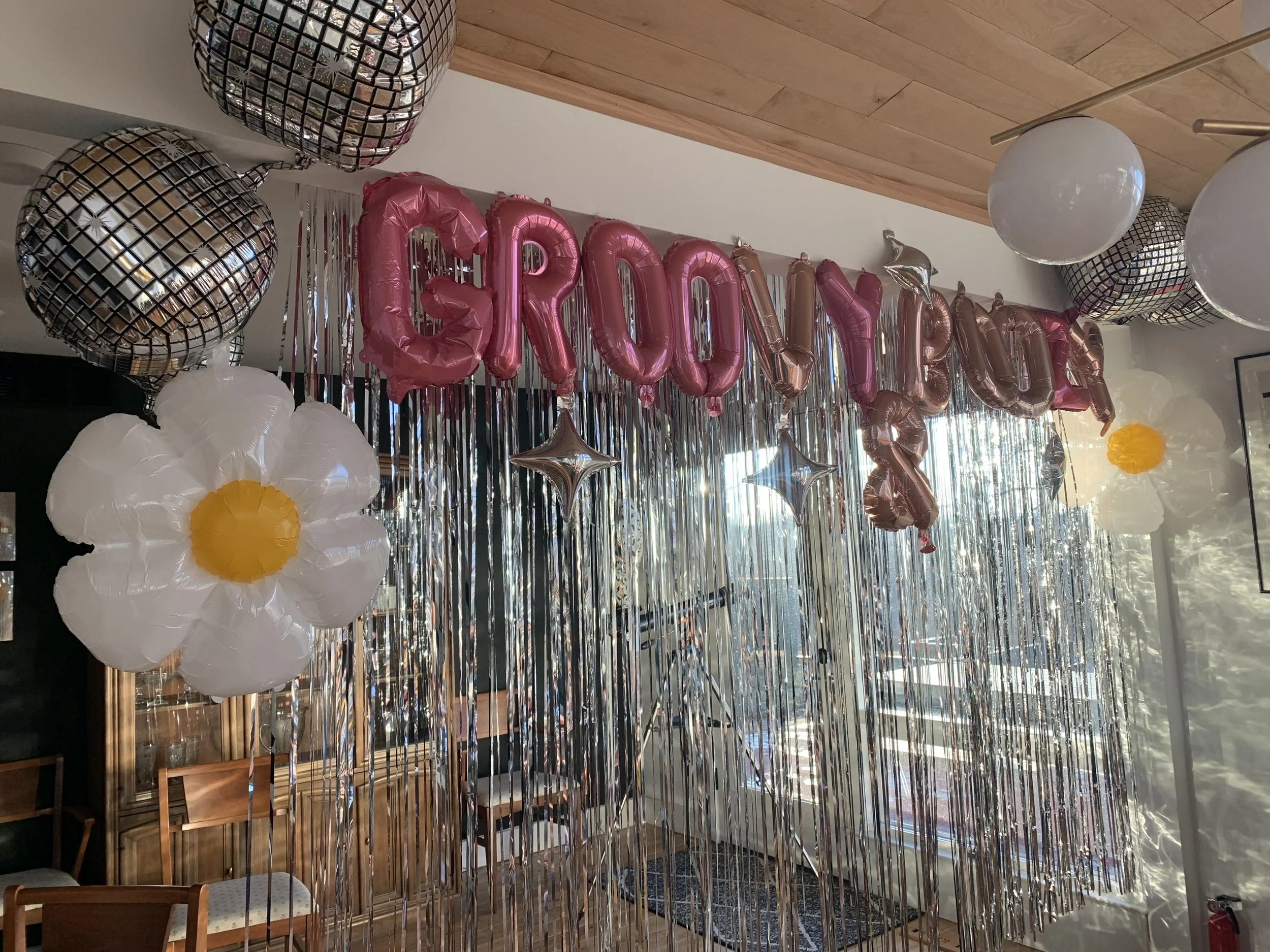 Decorative indoor scene with large metallic disco ball balloons, pink letter balloons spelling "GROOVY BIRTHDAY," white flower-shaped balloons with yellow centers, and hanging star balloons, with shiny silver streamers and a window in the background.