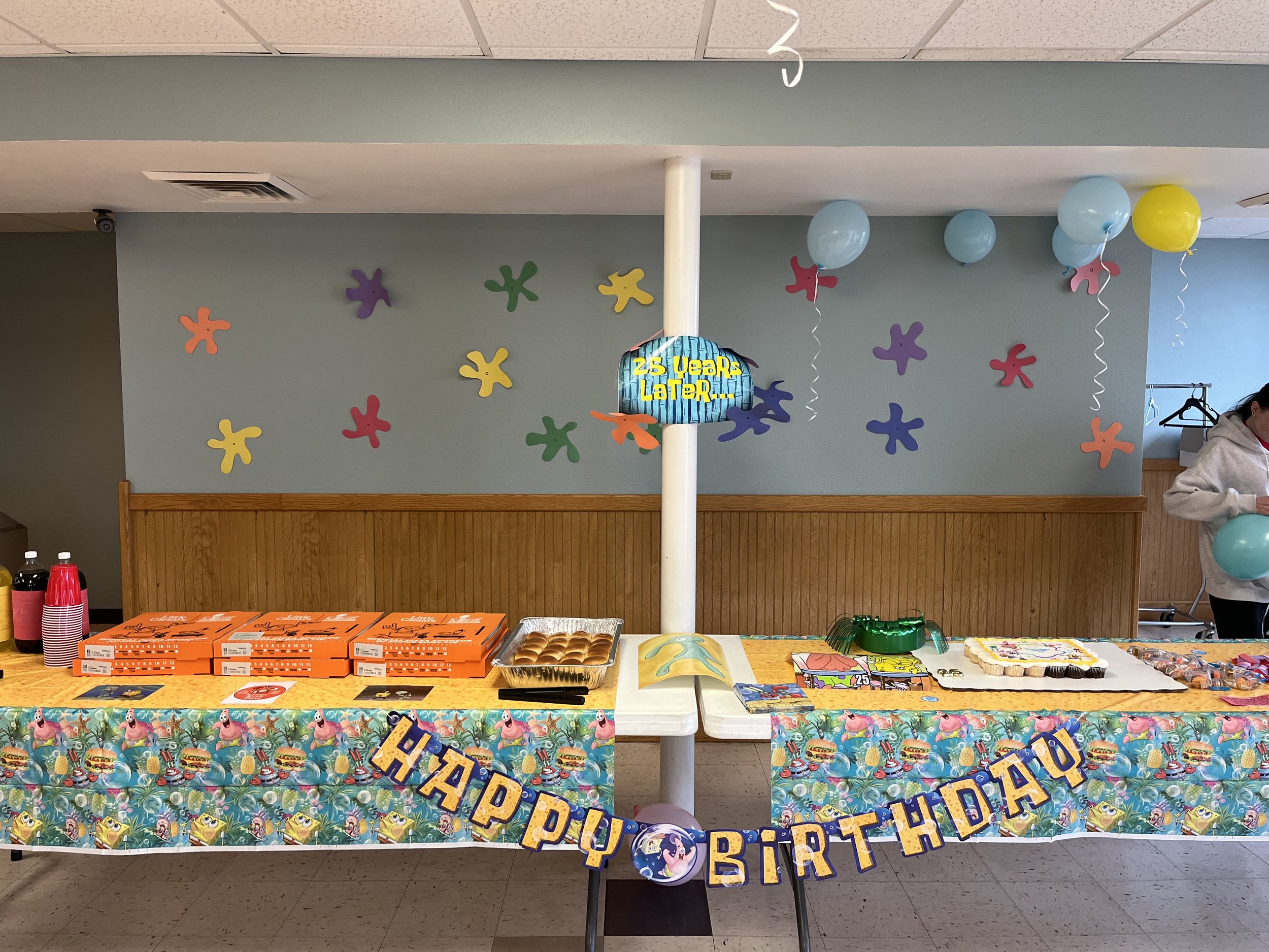 Party table decorated with a Hawaii-themed tablecloth, colorful balloons, and birthday banners. The table has pizza boxes, a cake, and party favors, with a sign celebrating 25 years.