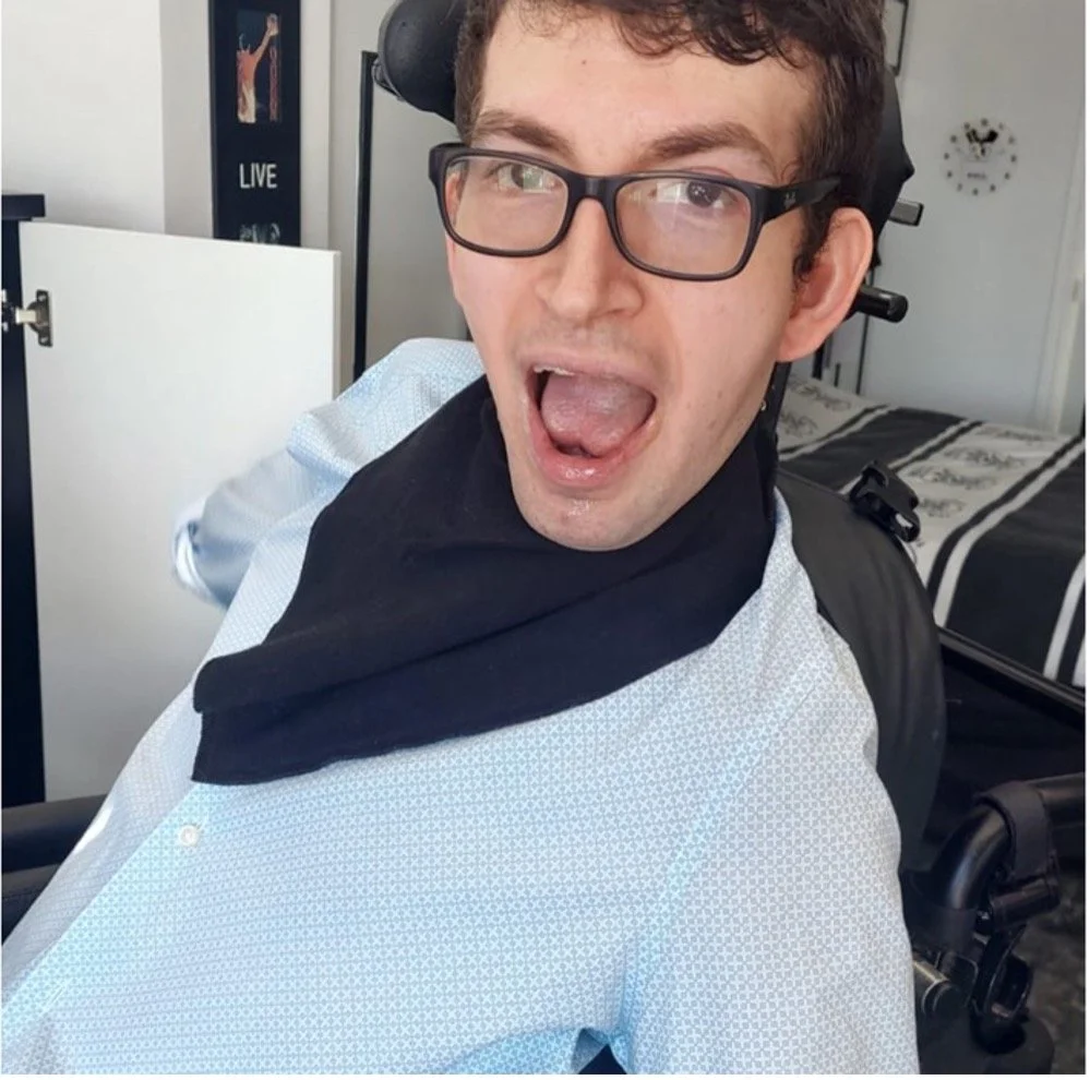 A young man with glasses, curly brown hair, and light skin, sitting in a wheelchair, making a surprised or excited expression with his mouth open, in a bedroom with a bed and a framed picture in the background.