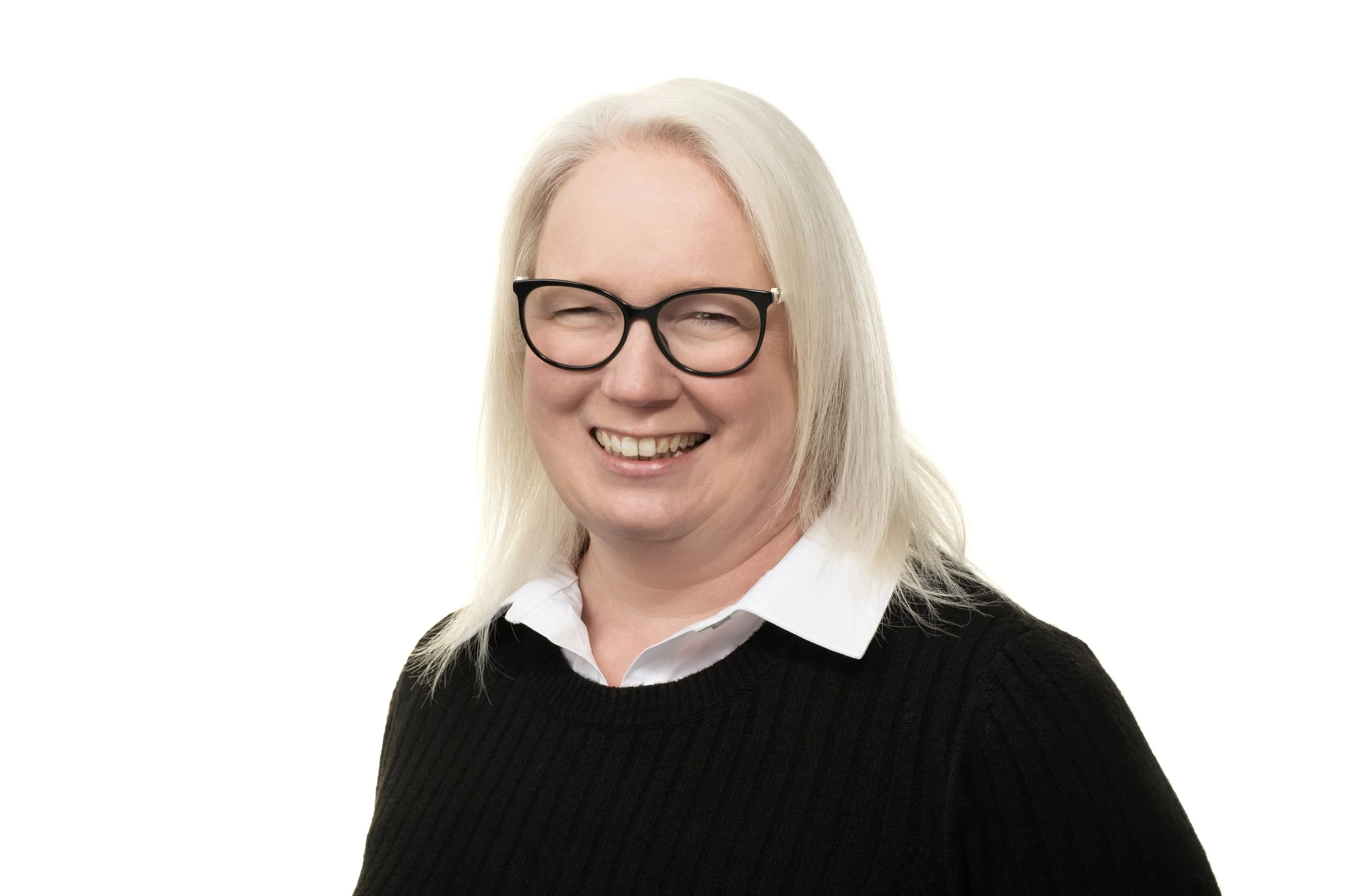 Portrait of a smiling woman with long blonde hair, black glasses, a white collared shirt, and a black sweater, against a plain white background.