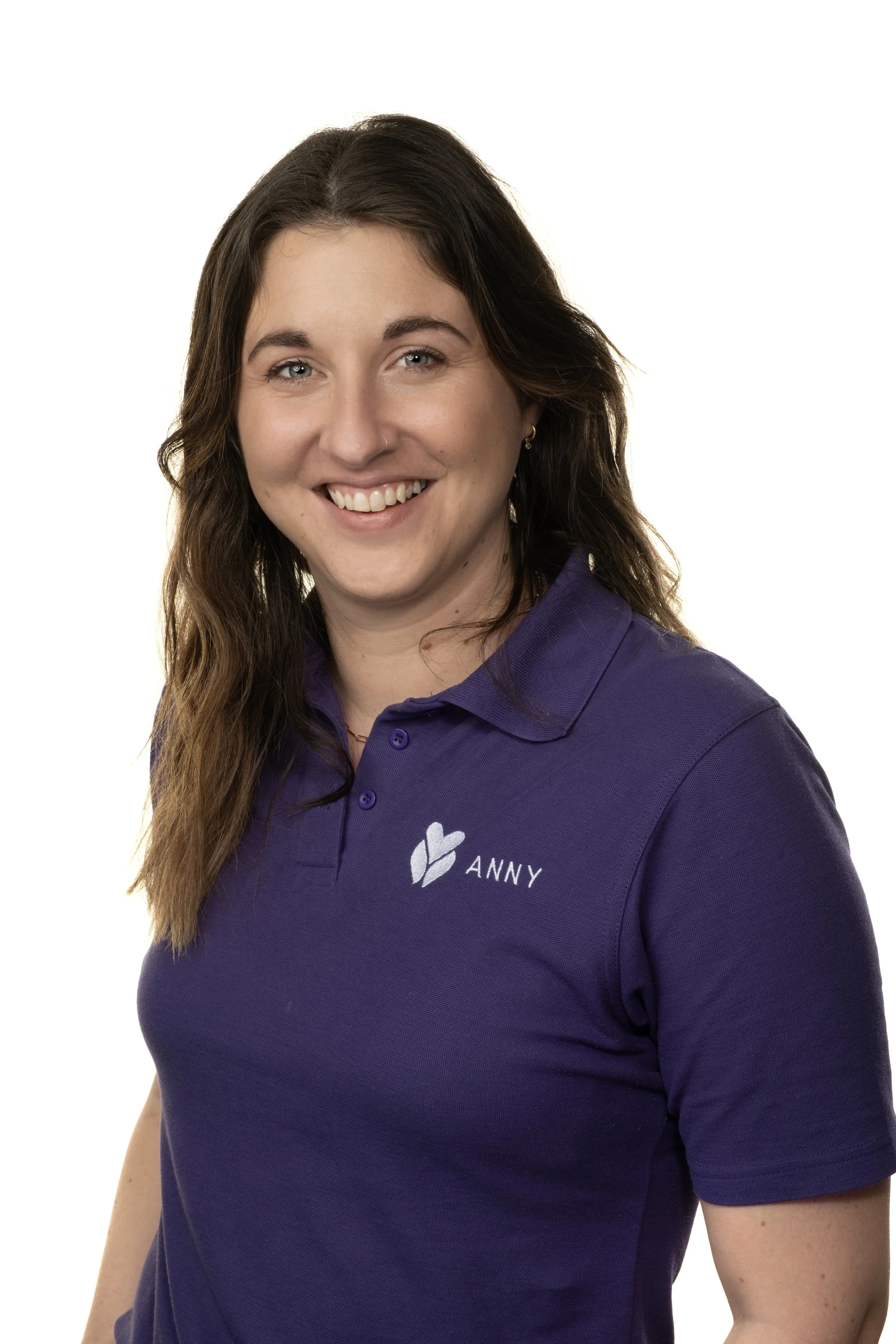 A woman with long brown hair wearing a purple polo shirt with a logo and the name 'Anny' embroidered on it, smiling at the camera with a white background.