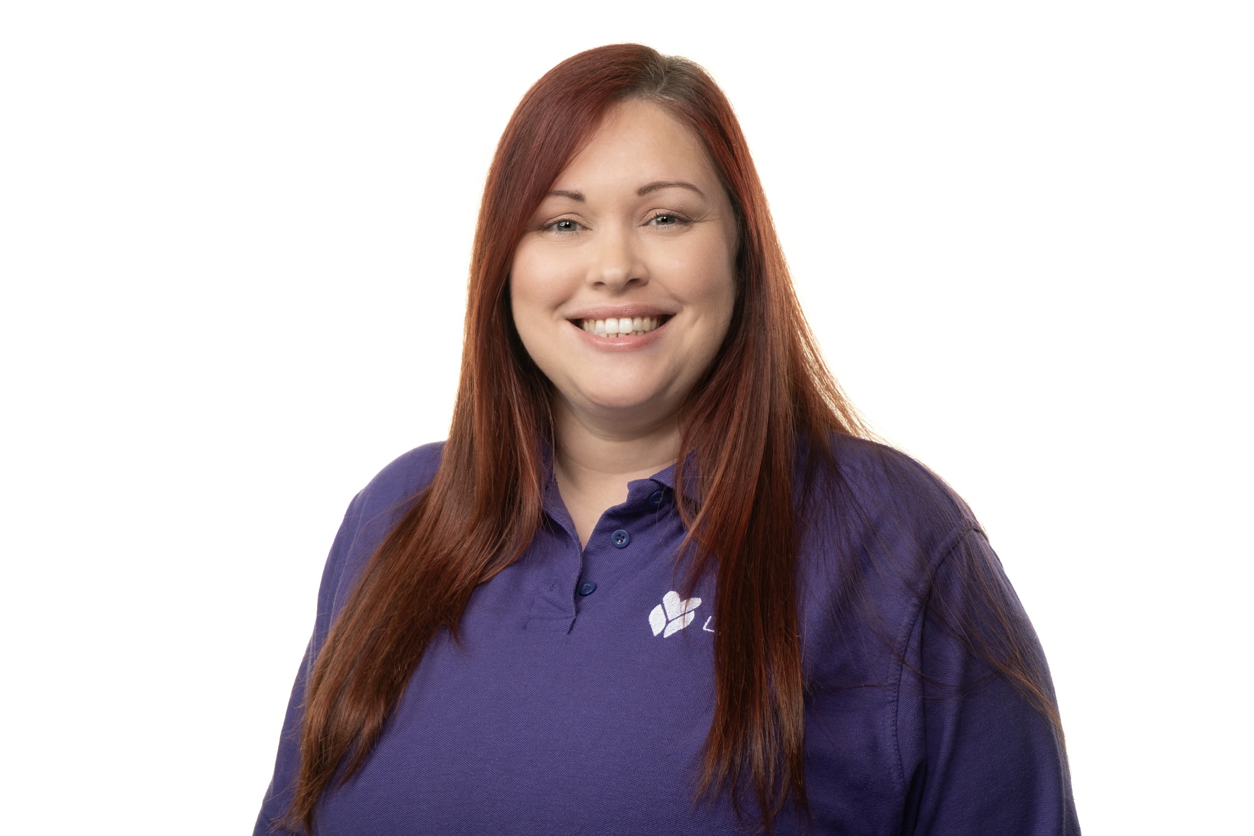A woman with long red hair smiling, wearing a purple polo shirt with a logo on the chest, against a plain white background.