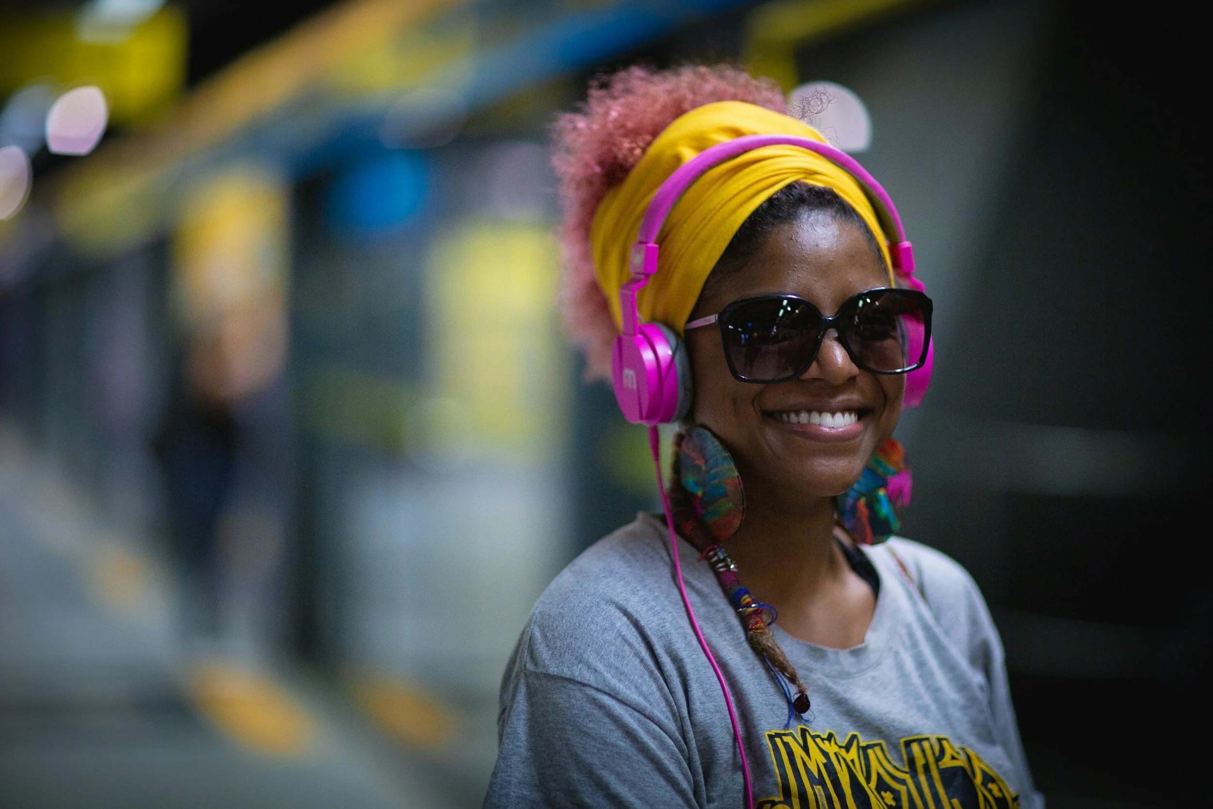 Portrait of a Black woman with black and pink hair, wearing headphones, sunglasses, a bright headband, and colourful earrings , smiling confidently in natural light