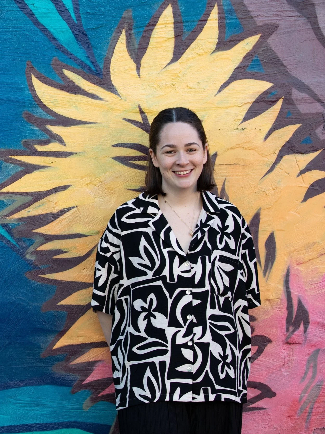 portrait photo of martha kennedy, neurodivergent affirming occuaptional therpist smiling in front of a wall of blue, pink and yellow street art