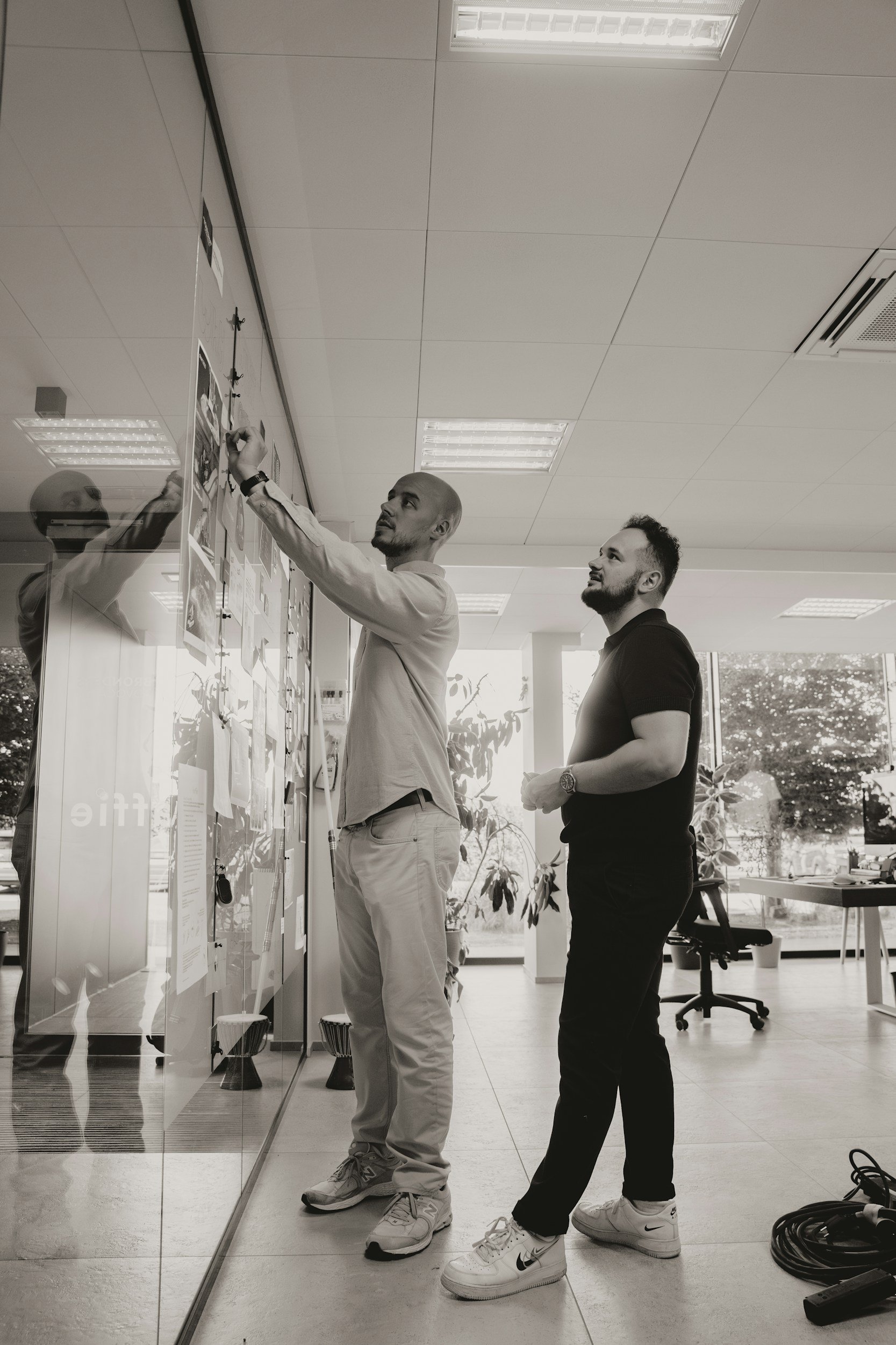 Deux hommes discutent devant un tableau blanc dans un bureau, l'un écrit ou colle quelque chose, l'autre observe. La scène est en noir et blanc.