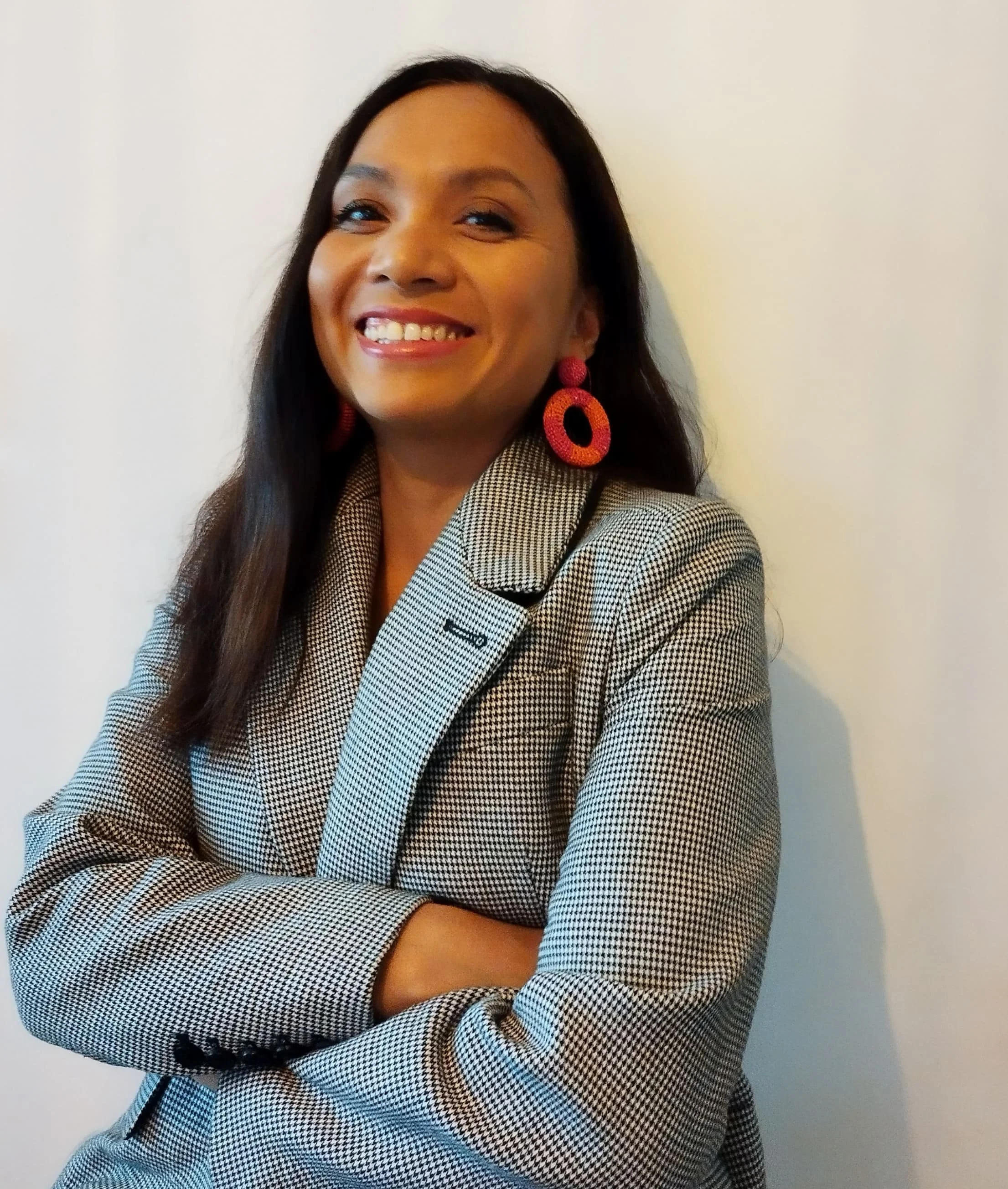 Femme souriante portant un blazer à carreaux et des boucles d'oreilles roses, bras croisés, contre un mur blanc.