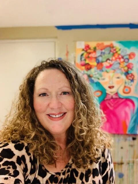 A smiling woman with curly hair taking a selfie in front of a colorful abstract painting of a woman with flowers in her hair.