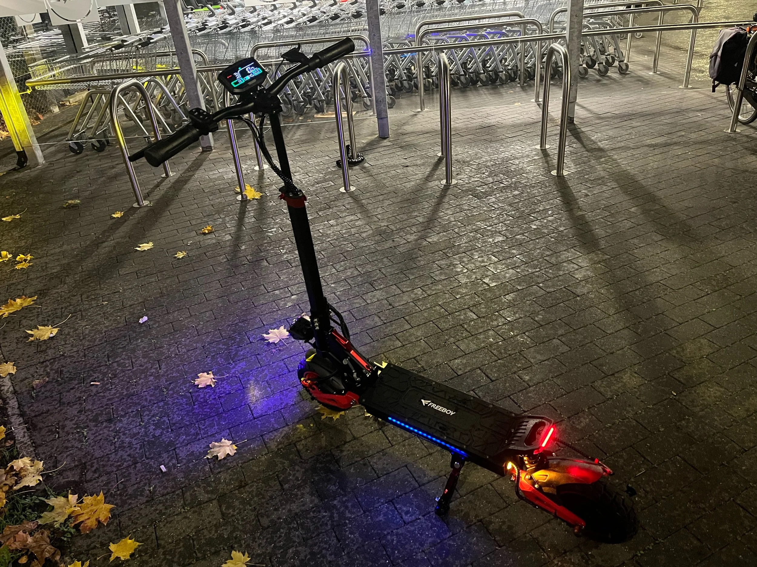 An electric scooter parked on wet pavement outside a building with bike racks and bicycles inside. The scooter has illuminated lights and a digital display on the handlebar.