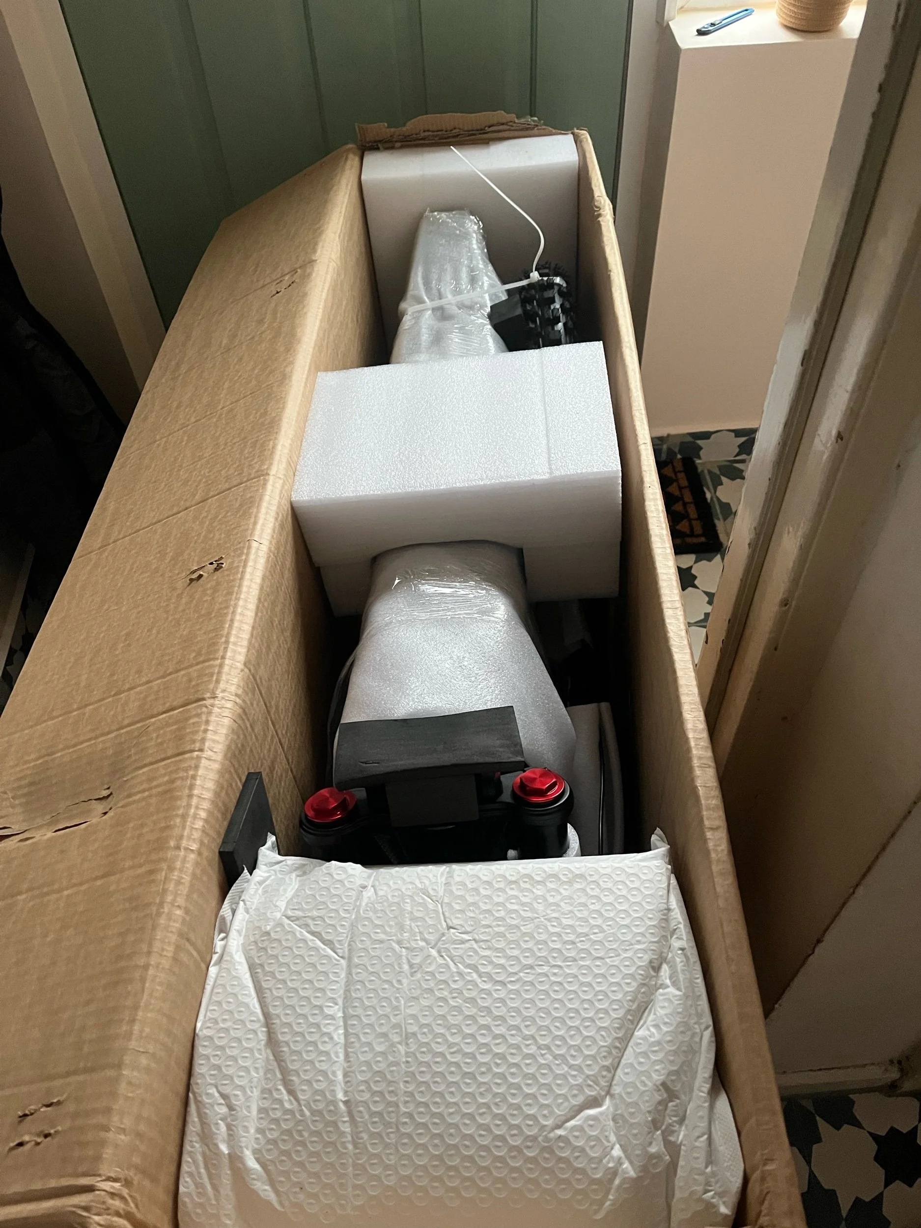 Unboxed scooter with wrapped handlebars and white foam packaging, placed inside a large cardboard box lined with brown paper, in a room with patterned floor tiles.