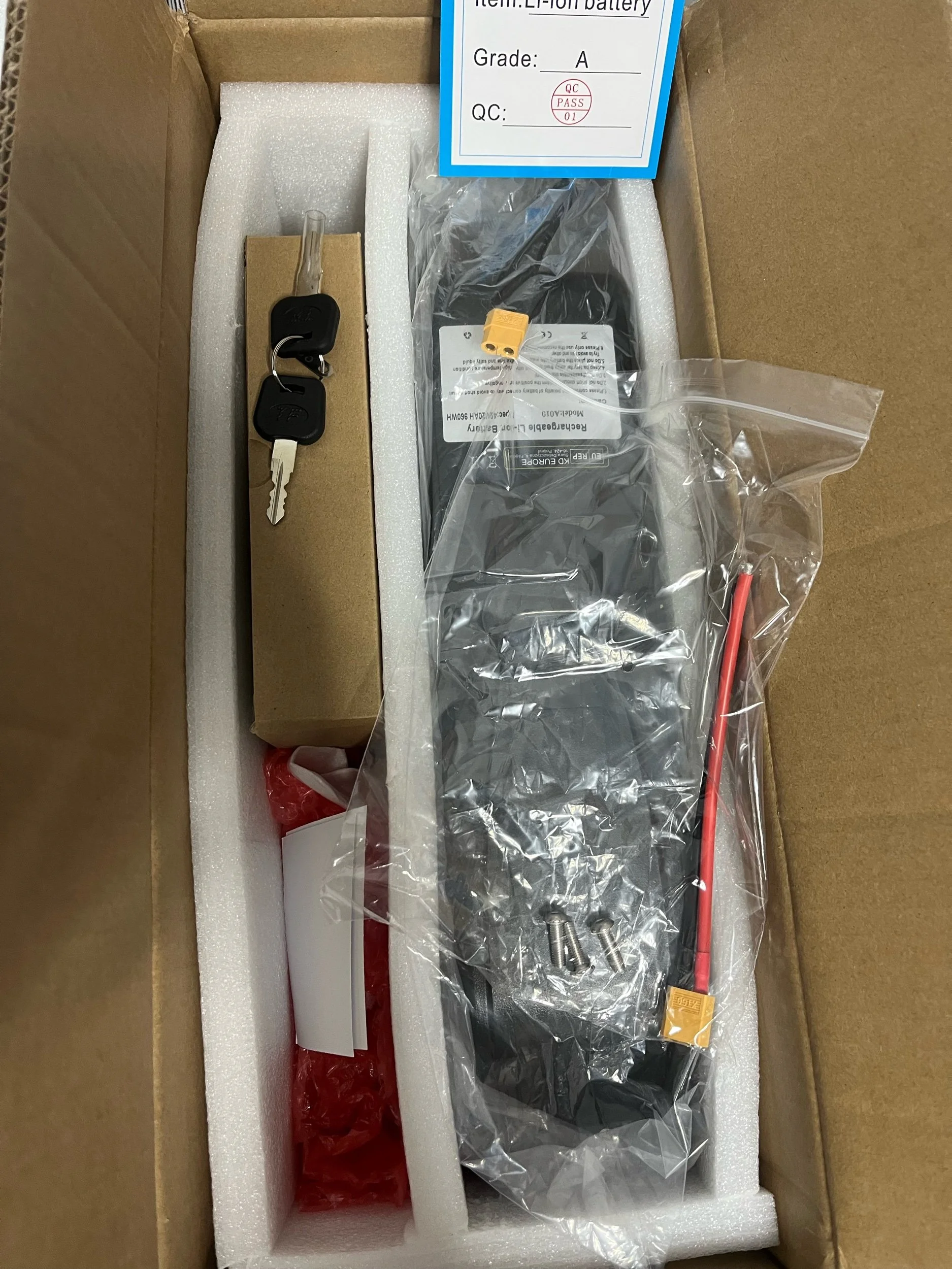Inside a cardboard box, there is a key with a black key fob, a black component in plastic packaging with a yellow connector, and additional small parts with red cable ties, all laid on white foam padding.