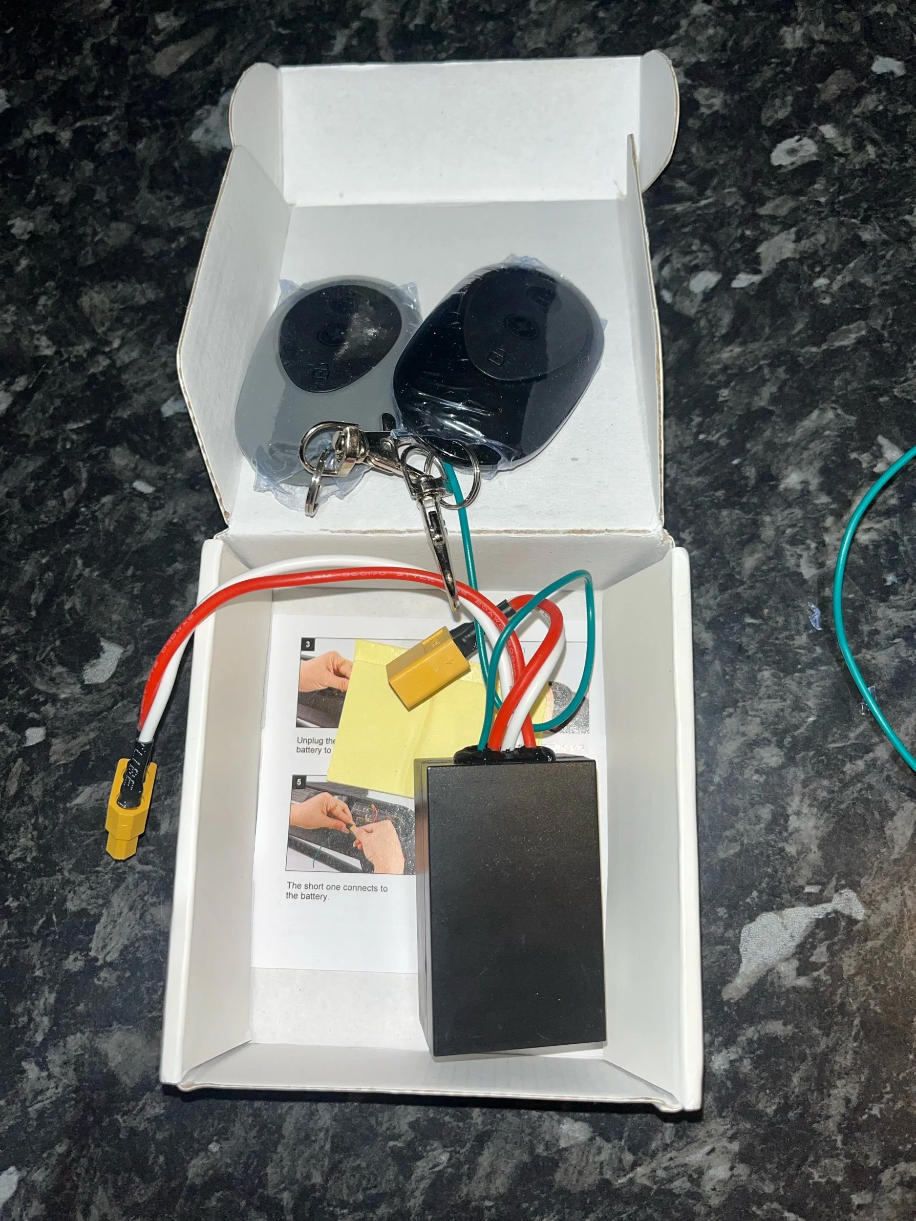 Open box containing two black remote controls, a black rectangular device, and various colored wires connected to the devices, with instructions printed inside.