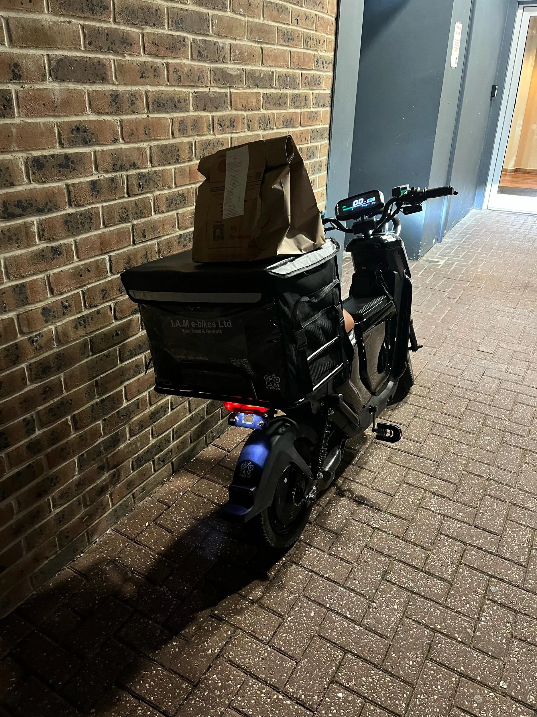 An electric bicycle with a large cargo bag and a paper bag on top, parked against a brick wall near a doorway.