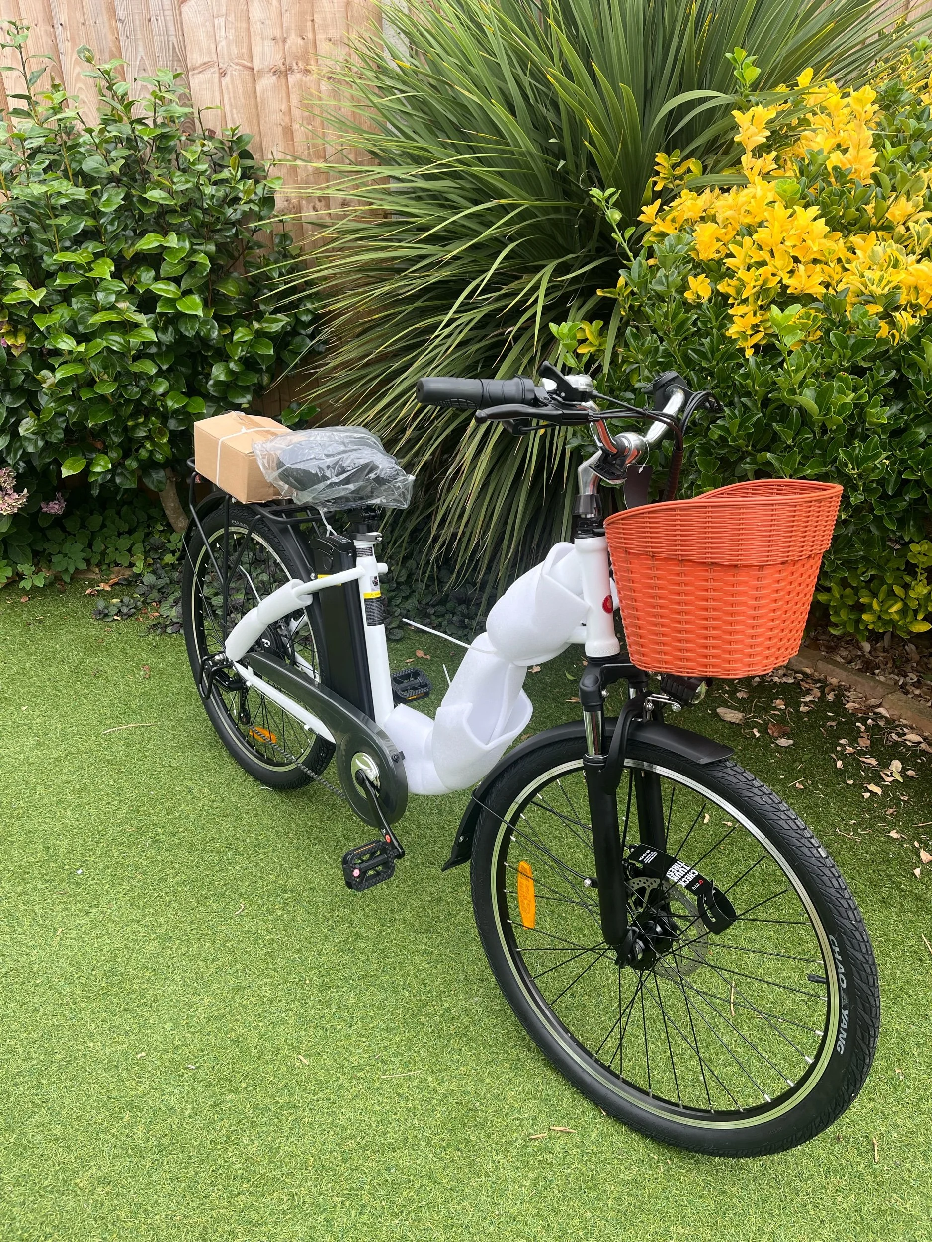 A white electric bicycle with black accents and an orange front basket, parked on a grassy area with lush green bushes and yellow flowers in the background.