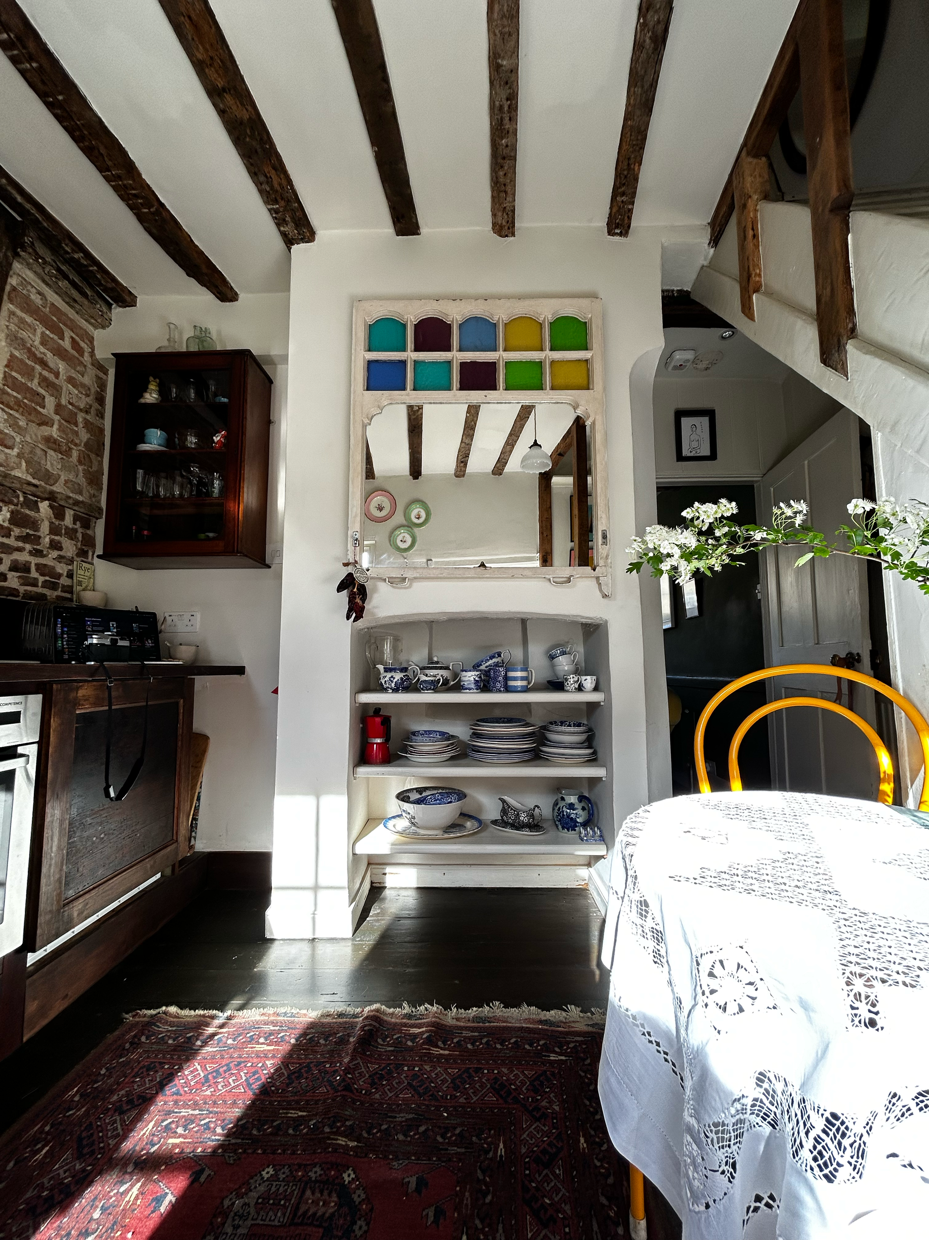 A cottage beamed ceiling kitchen. Spode mixed crockery, reclaimed mirror, vintage rug and chairs and antique linen table cloth on a small round table 