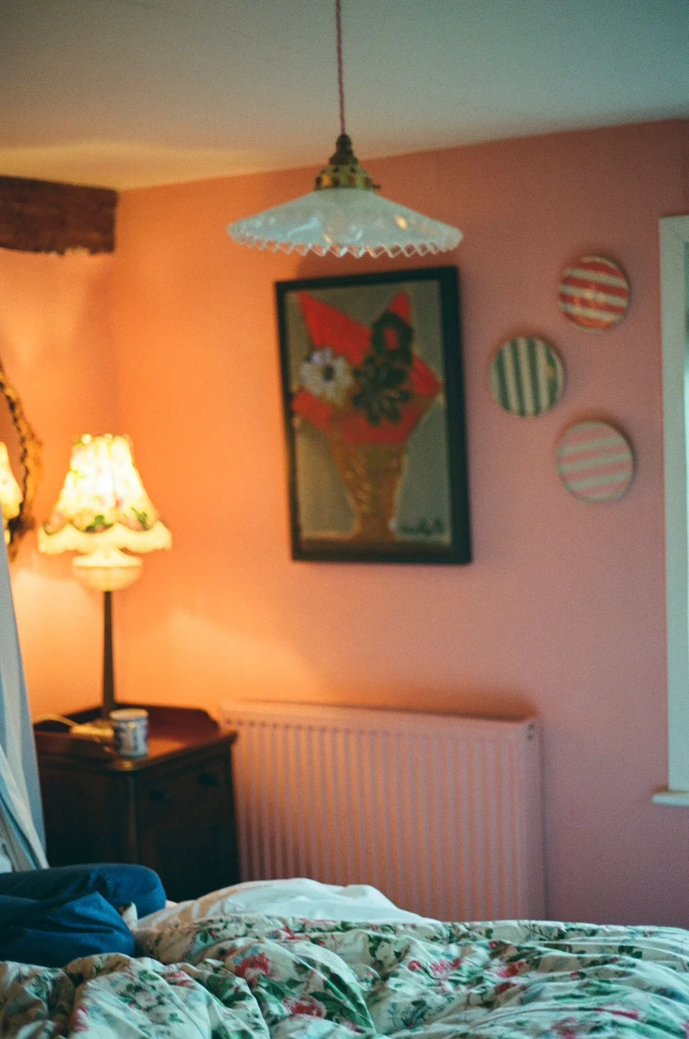 A whimsical cottage bedroom with Farrow and Ball Blooth Pink wall, a Luke Hannam painting and Kate Sellers plates on the wall, a lamp with a Sanderson lampshade. Nora Ephron vibes.