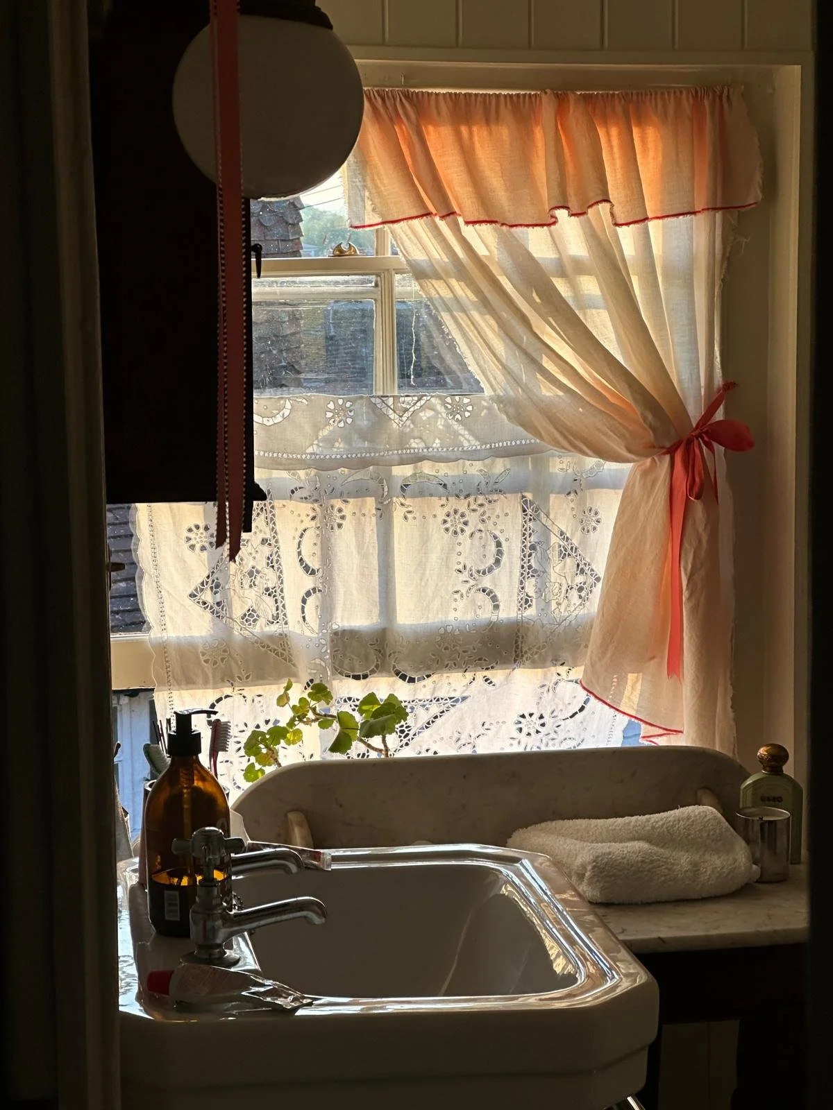 Whimsical bathroom window at Dot Cottage Rye with a peach ribbon- tied curtain and vintage table cloth detail, creating a charming storybook cottage atmosphere