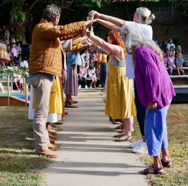 People standing in two lines facing each other, holding hands and dancing outdoors at a gathering or festival with seating and trees in the background.