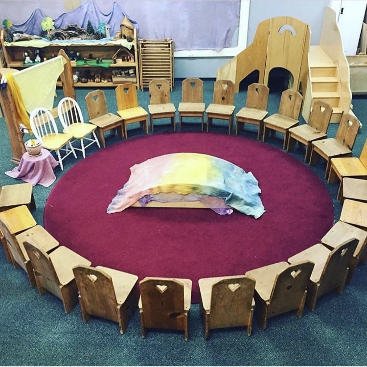 A circle of wooden chairs arranged on a red circular rug with a colorful cloth draped over it in the center. The chairs are in a room with a green carpet and a bookshelf or storage unit in the background. The room appears to be set up for a children'