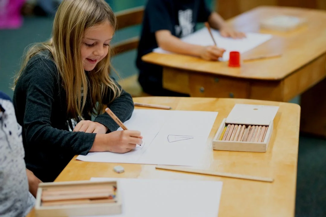 A young girl is drawing on a piece of paper at a classroom desk, smiling, with boxes of colored pencils nearby.