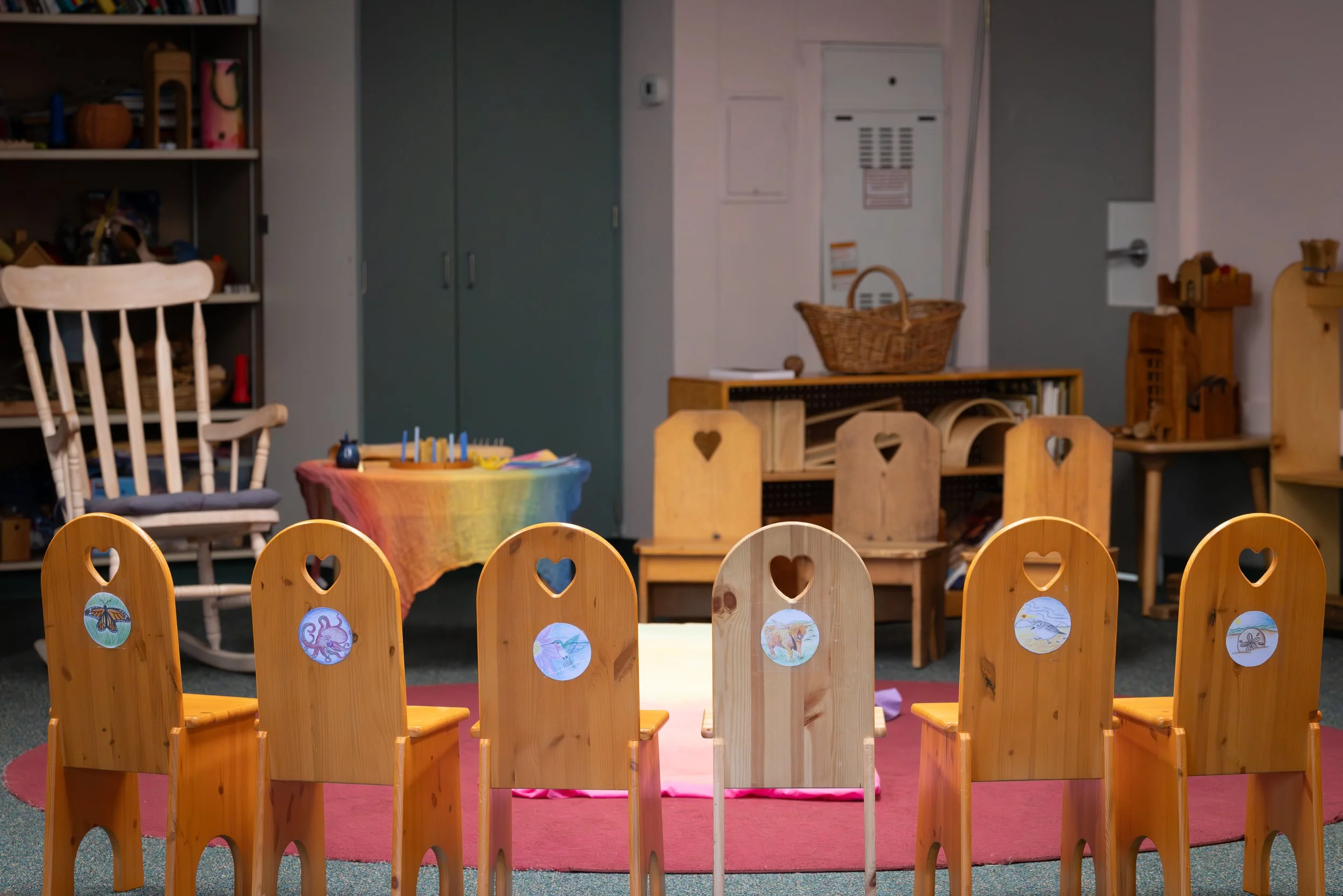 A circle of wooden chairs with heart-shaped cutouts on the backrests, each decorated with animal stickers, around a pink rug in a room that appears to be a classroom or play area.