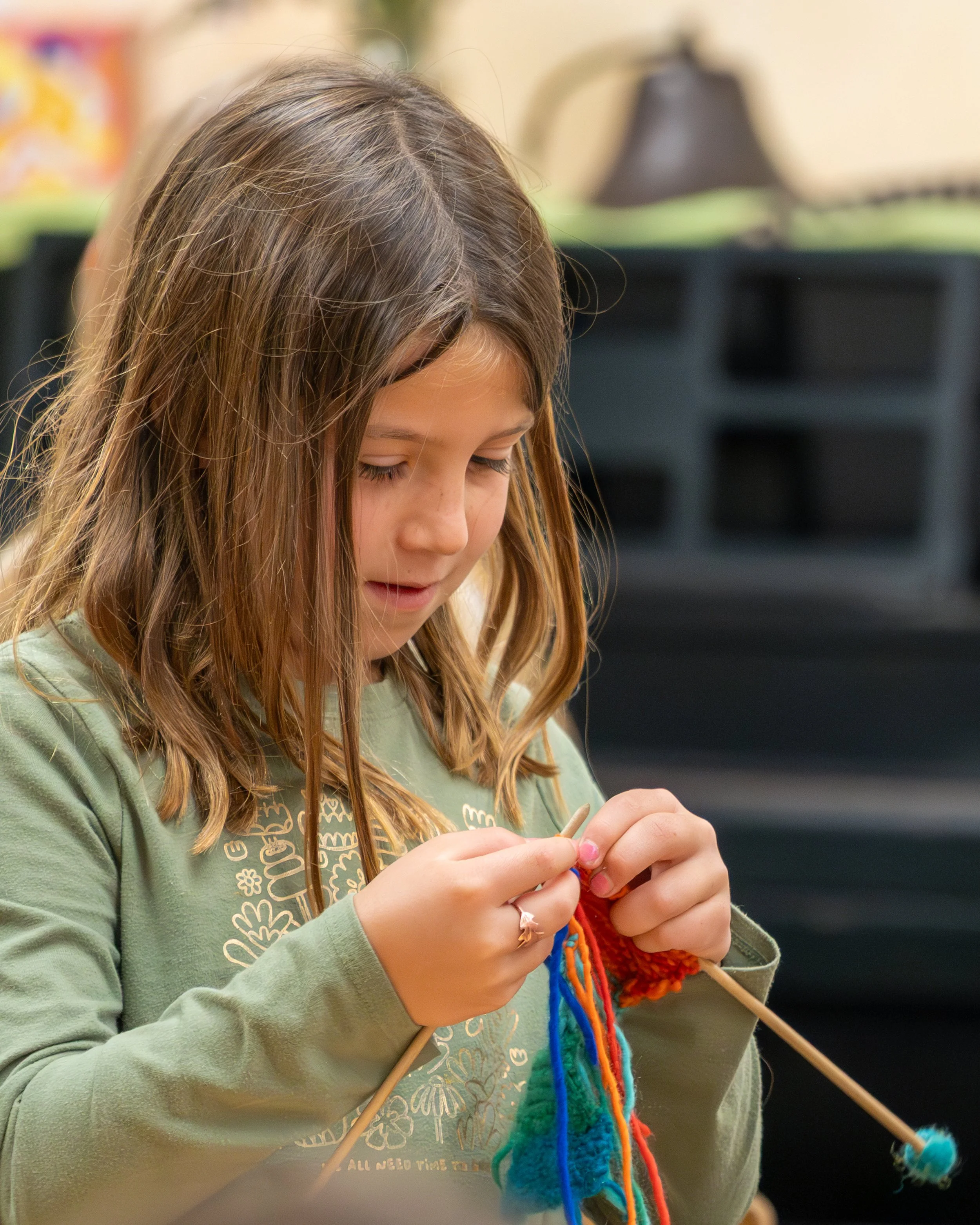 A young girl knitting with colorful yarn and wooden knitting needles in an indoor setting.