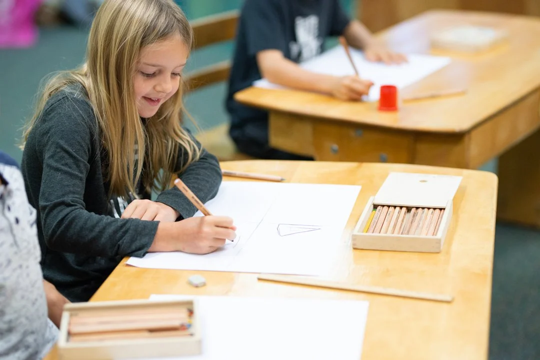 A young girl sitting at a wooden desk, drawing with a brown crayon on a sheet of paper, with other children in the background.